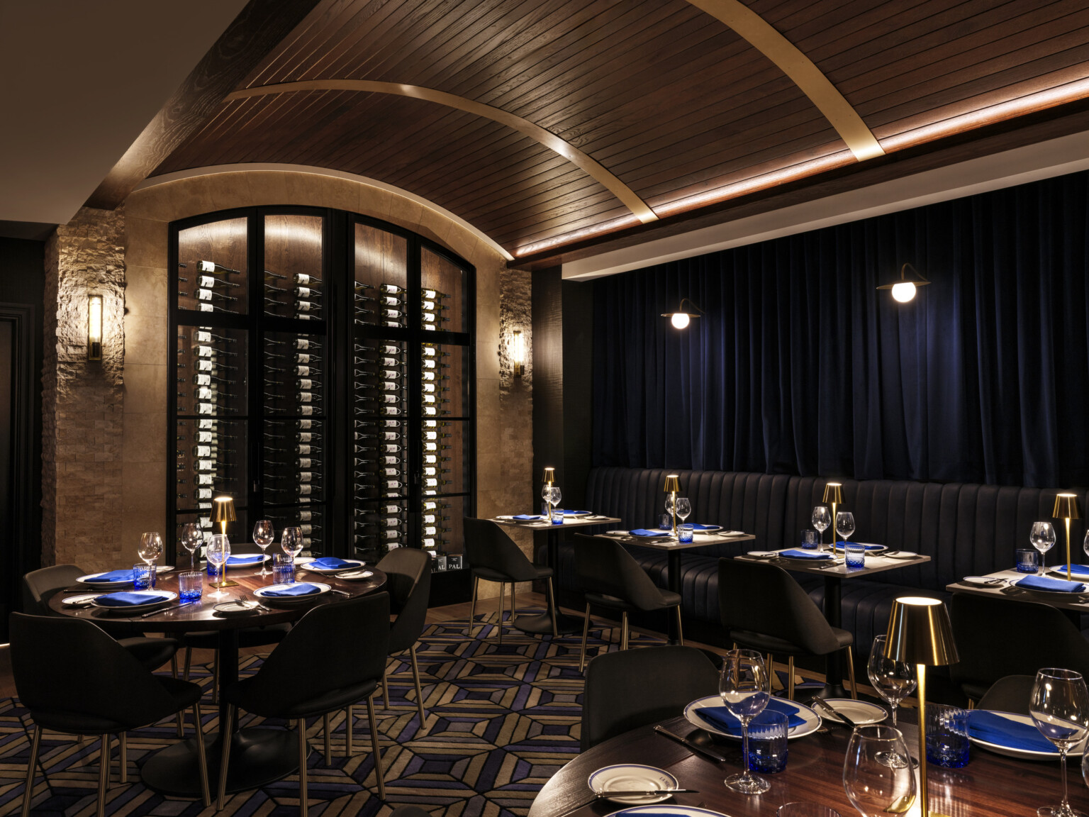 dining space in The Vanguard Hotel, Autograph Collection hotel; dimly lit moody cocktail lounge type space with dark wood table and chairs throughout; blue table decorations are lit by smaller lamplights; arched accent wall to the back of the room with wine bottled stacked behind glass doors