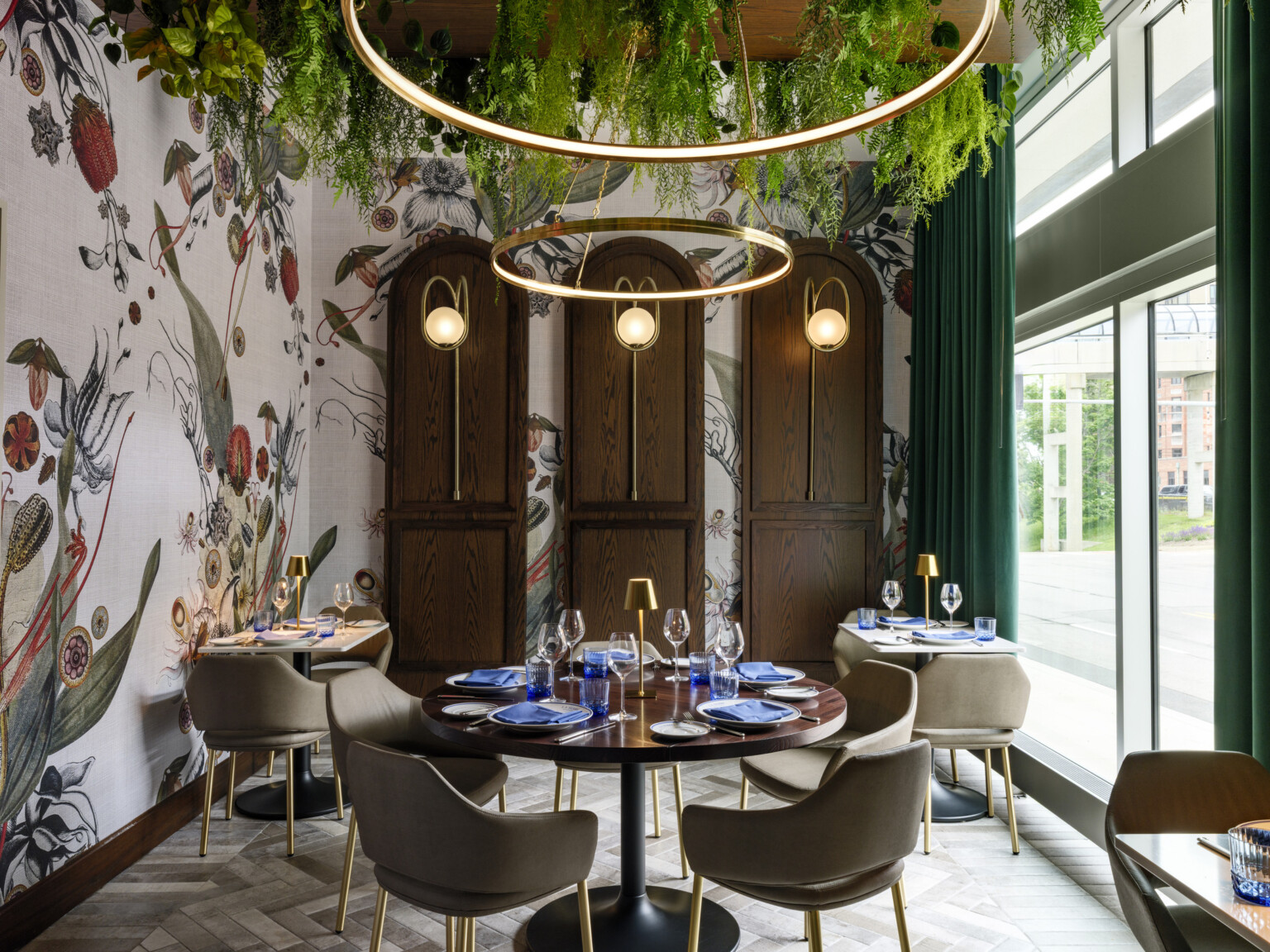 dining space in the The Vanguard Hotel, Autograph Collection hotel; greenery hangs from ceiling and integrates with the circular lighting fixtures; looking down on small ornately decorated tables with blue accents; floor to ceiling windows on the right wall