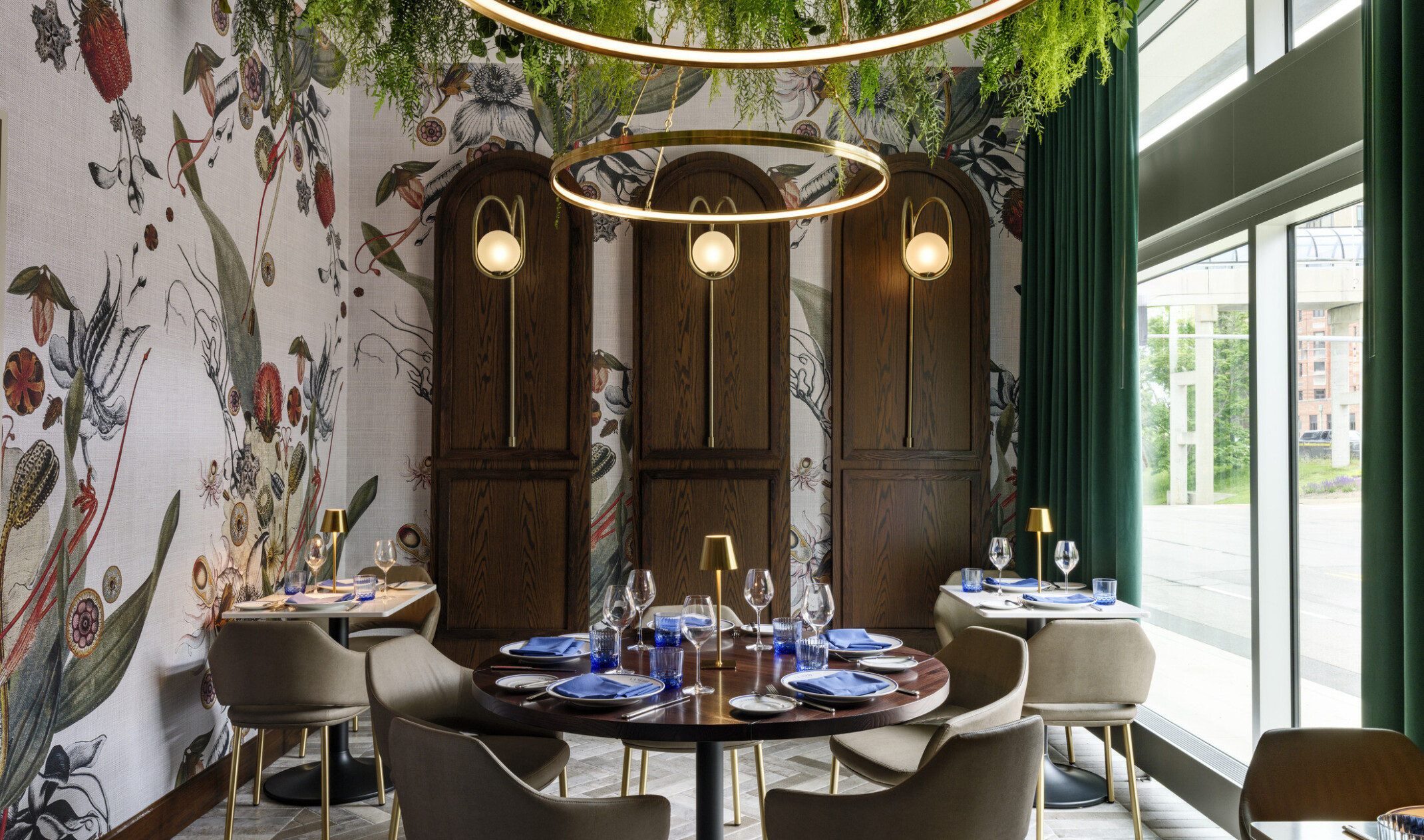 dining space in the The Vanguard Hotel, Autograph Collection hotel; greenery hangs from ceiling and integrates with the circular lighting fixtures; looking down on small ornately decorated tables with blue accents; floor to ceiling windows on the right wall