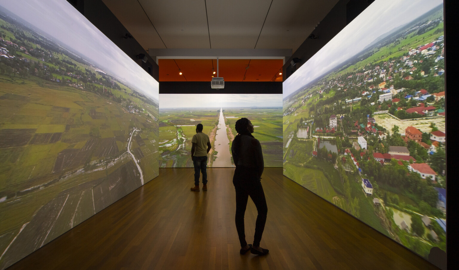 two darkened silhouettes stand in room with three walls with aerial images of farm lands, houses, and a river projected