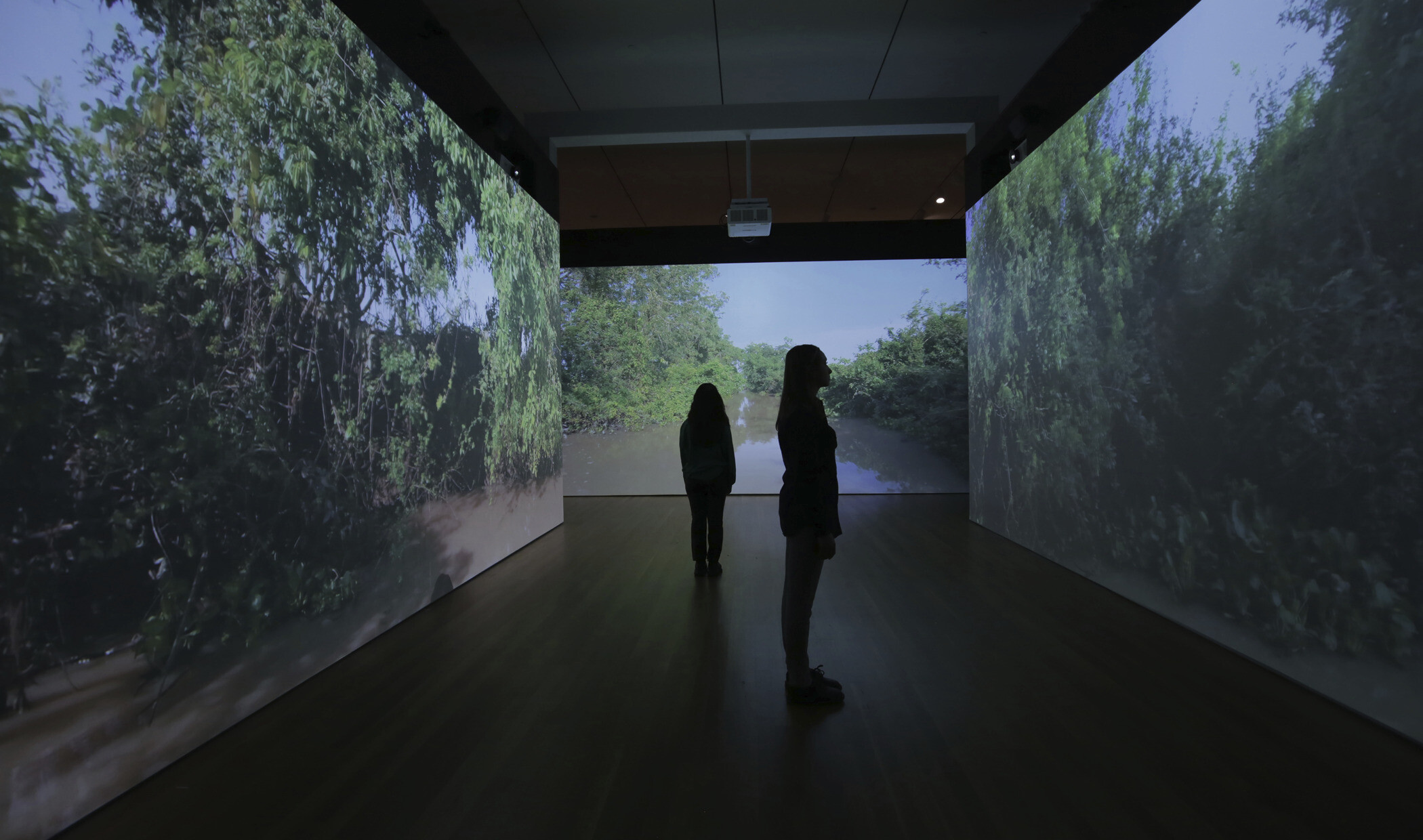 two darkened silhouettes stand apart in a dark room with three walls illuminated by projected images of a muddy river