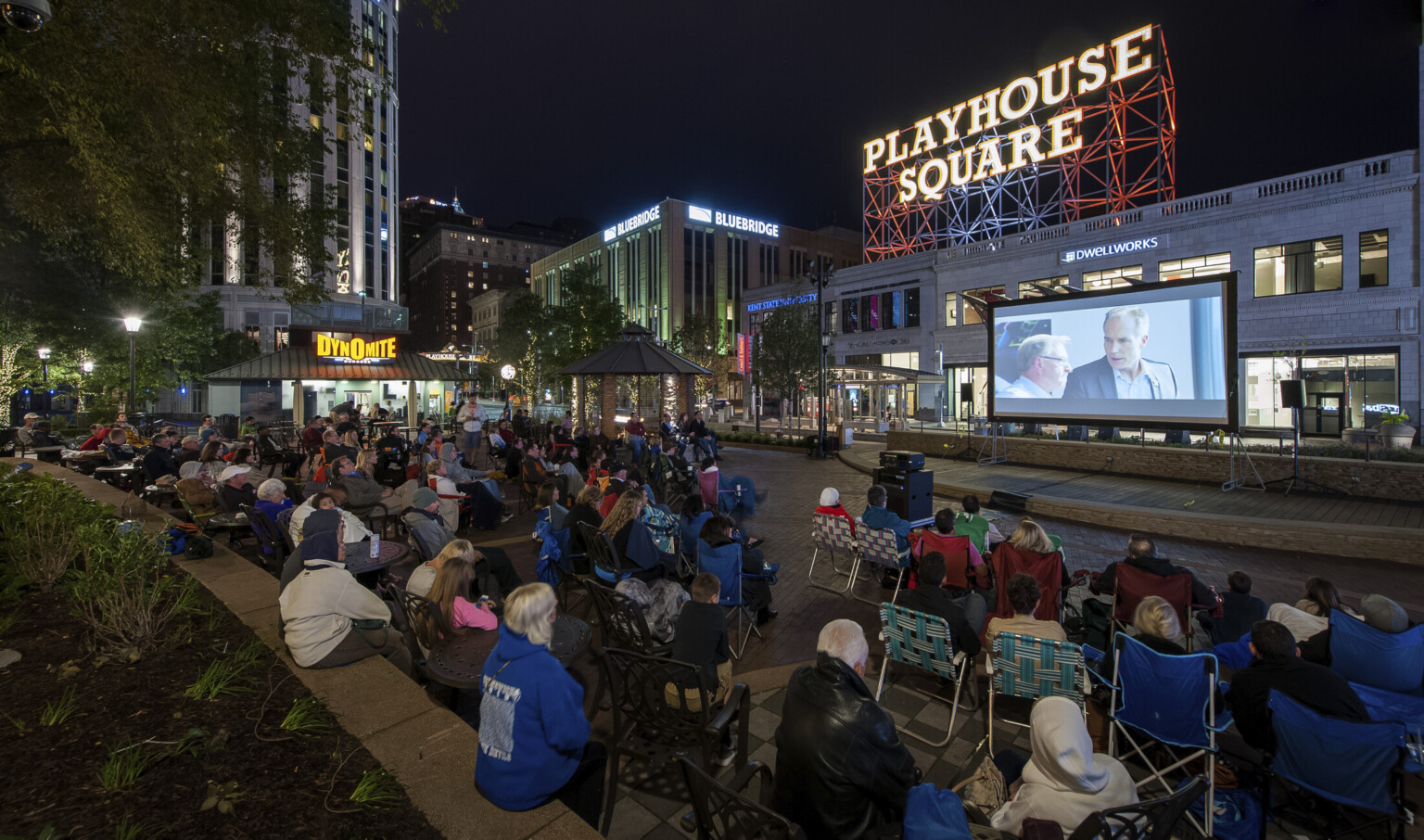 Audience sits on the street watching an outdoor movie on a screen. A Playhouse Square sign is illuminated in the background