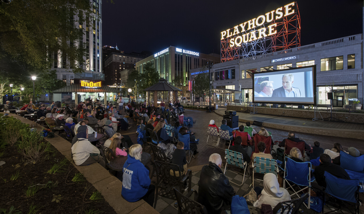 Audience sits on the street watching an outdoor movie on a screen. A Playhouse Square sign is illuminated in the background