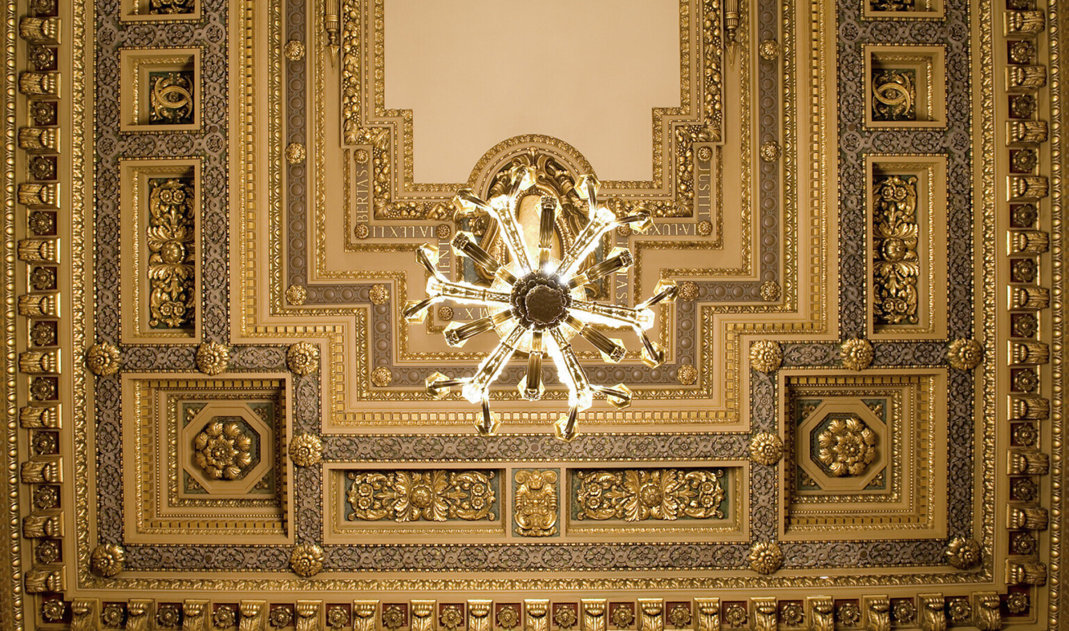 Looking up at ornate orthogonal ceiling detailed with floral medallions, roman phrases, cornice edging, and offset chandelier Looking up at ornate orthogonal ceiling detailed with floral medallions, roman phrases, cornice edging, and offset chandelier