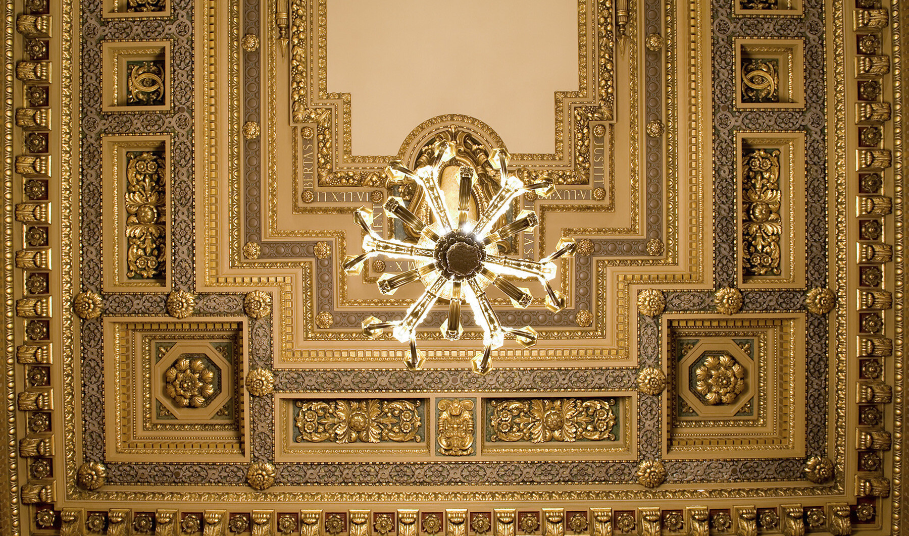 Looking up at ornate orthogonal ceiling detailed with floral medallions, roman phrases, cornice edging, and offset chandelier