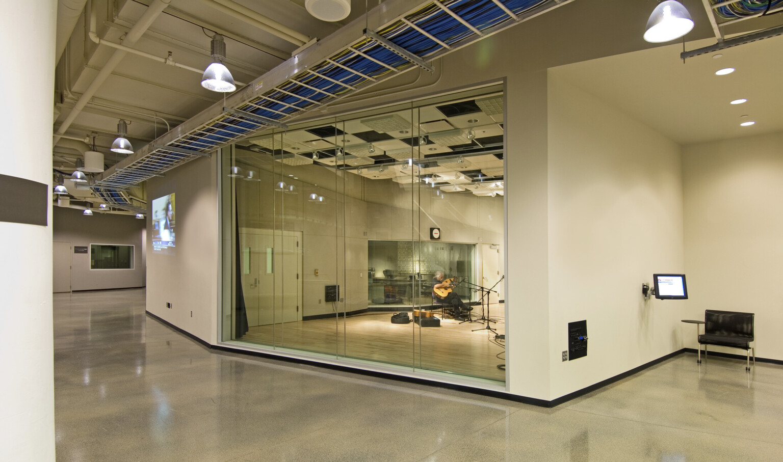 Guitarist in room with partial glass walls, hard wood flooring, and acoustic panels on ceiling. Outside room is a hall.