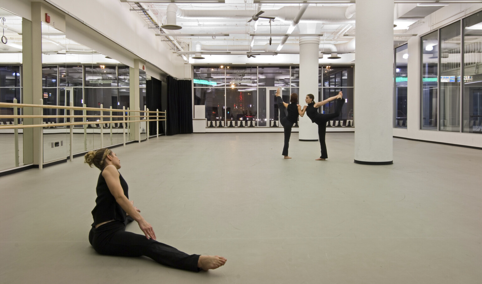 Three dancers in a white studio with large window walls looking out in the city. Left wall is mirrored and has a barre.
