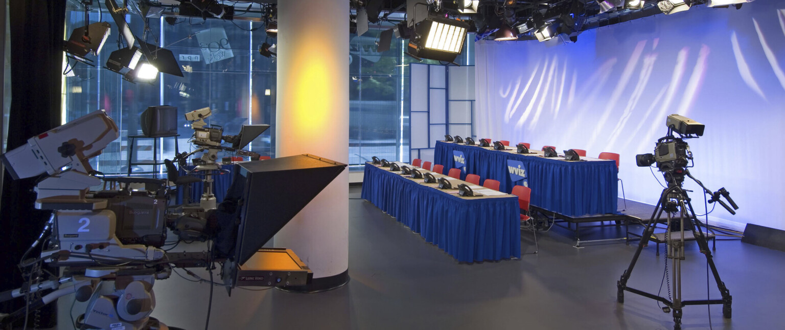 Television studio at street level with three cameras pointed at two rows of tables with chairs and phones, lit from above