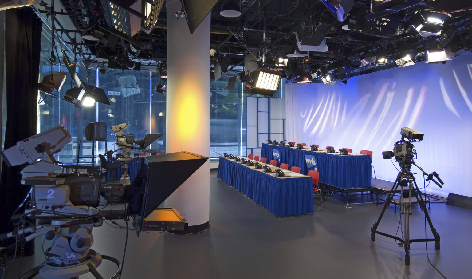 Television studio at street level with three cameras pointed at two rows of tables with chairs and phones, lit from above