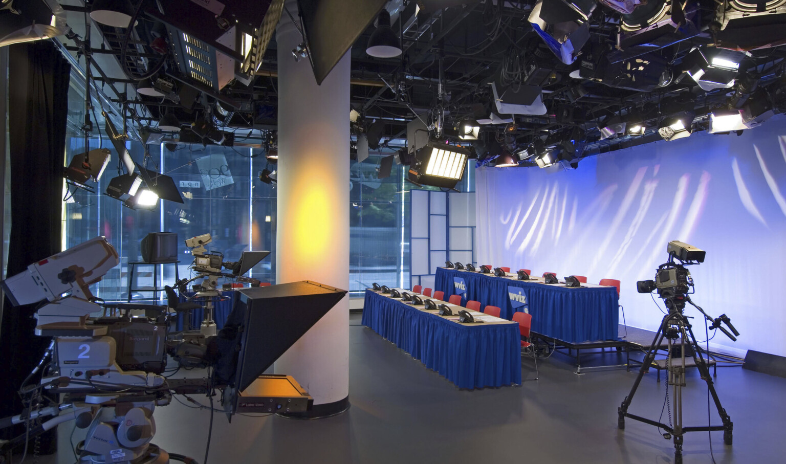 Television studio at street level with three cameras pointed at two rows of tables with chairs and phones, lit from above
