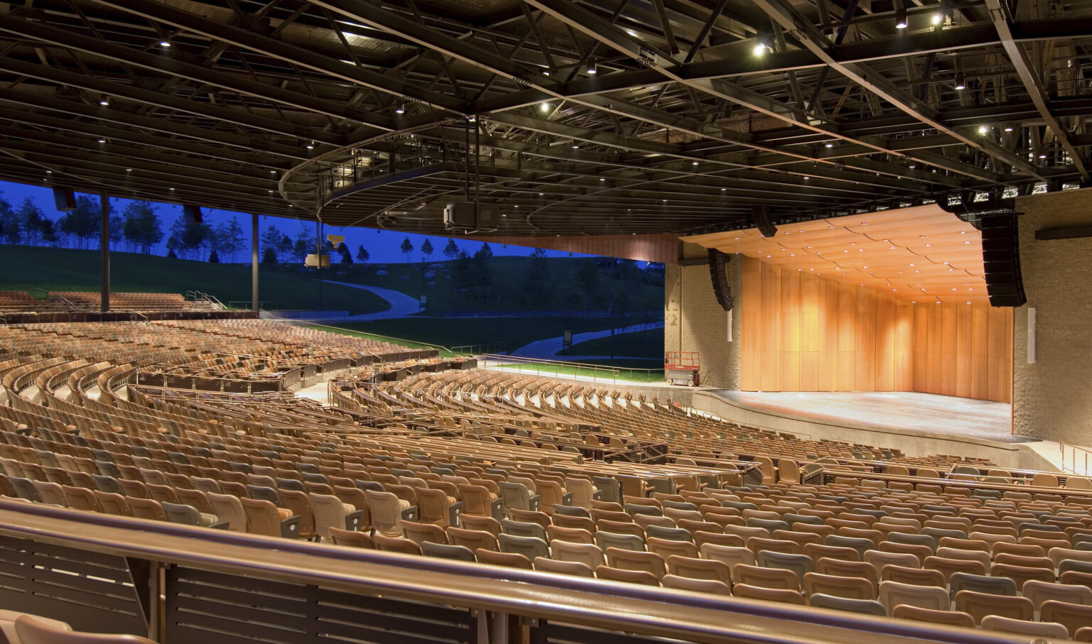 Light brown seats under black amphitheater roof facing wood paneled stage with rounded panel ceiling and recessed lights