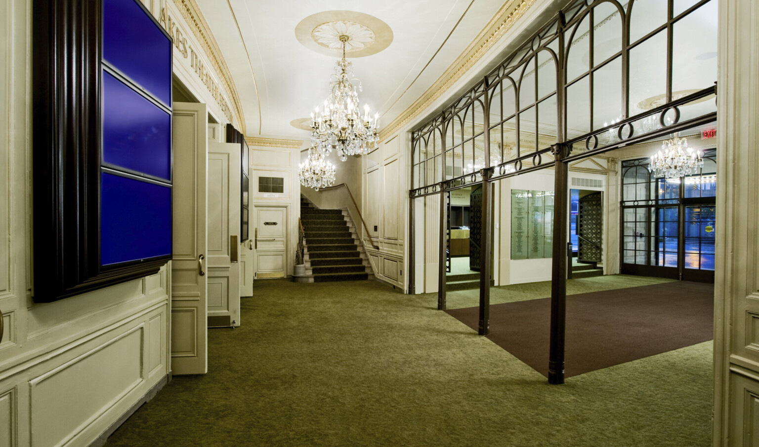 Green carpeted hallway leading to stairs, white walls with doors left and chandeliers hang from above framed by gold molding