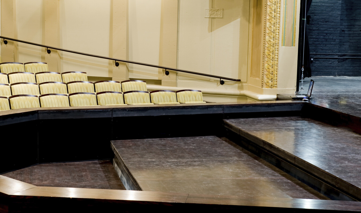 Thrust style proscenium with three black steps leading up to the stage in front of cream colored audience seats
