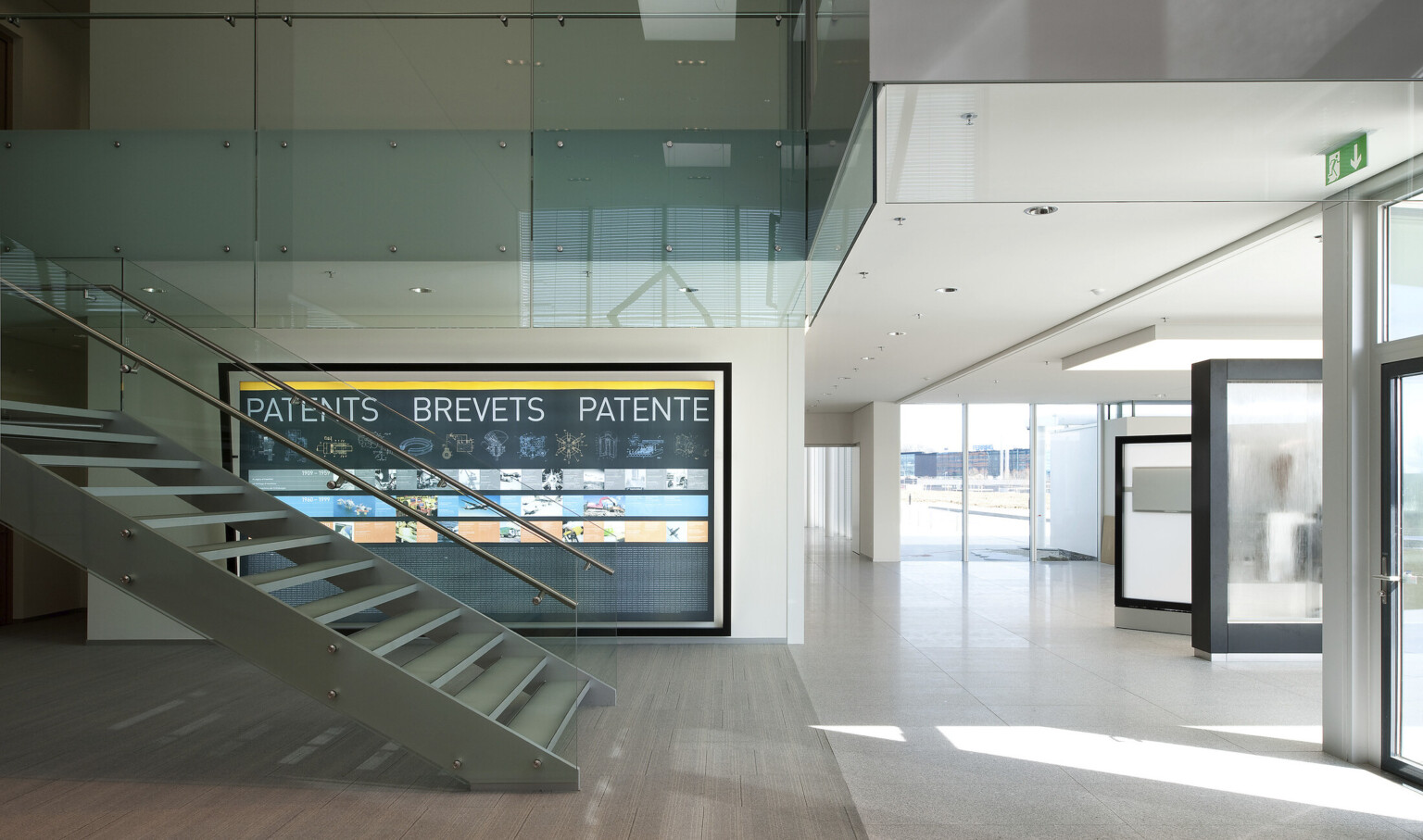 White hallway with floor to ceiling windows, right. Floating stairs with glass bannister, left. Back, infographic of patents