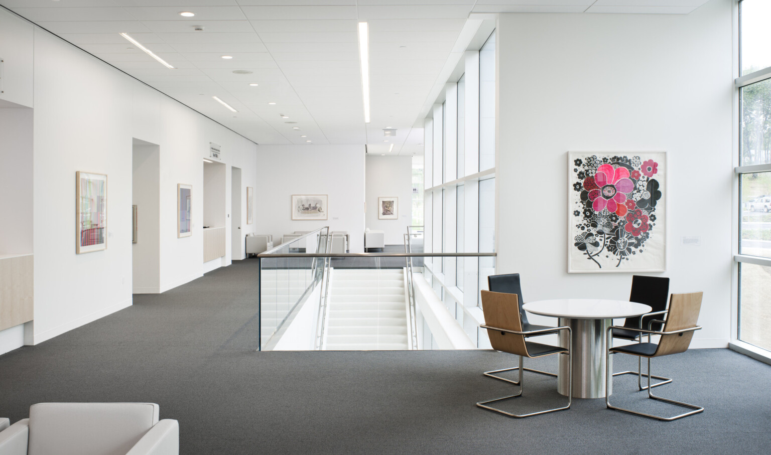 White room with multiple seating areas and stair case center coming from below next to window wall. Art is framed on wall