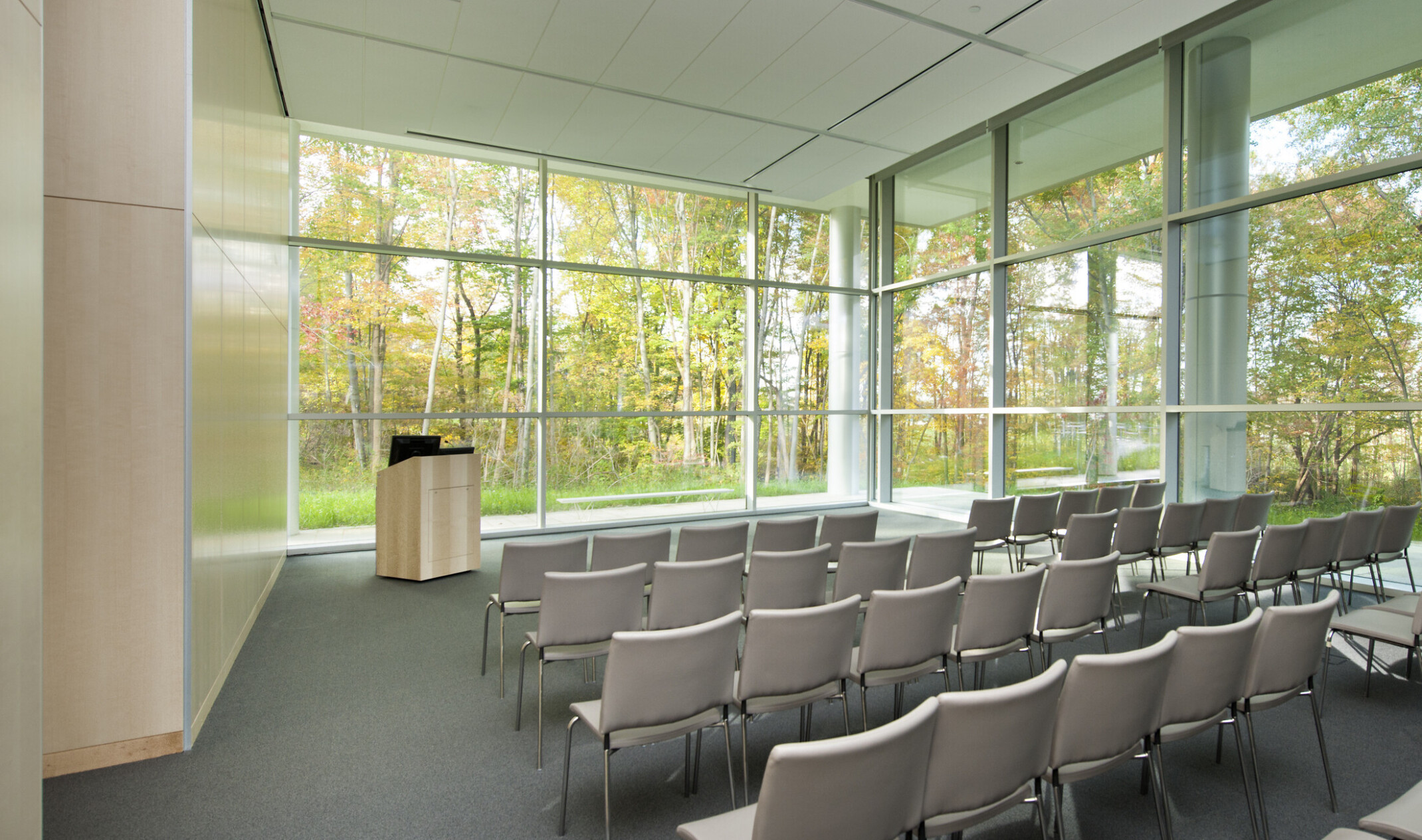 Auditorium with grey floor, wood left wall, and floor to ceiling windows on two walls with ground level view of trees.