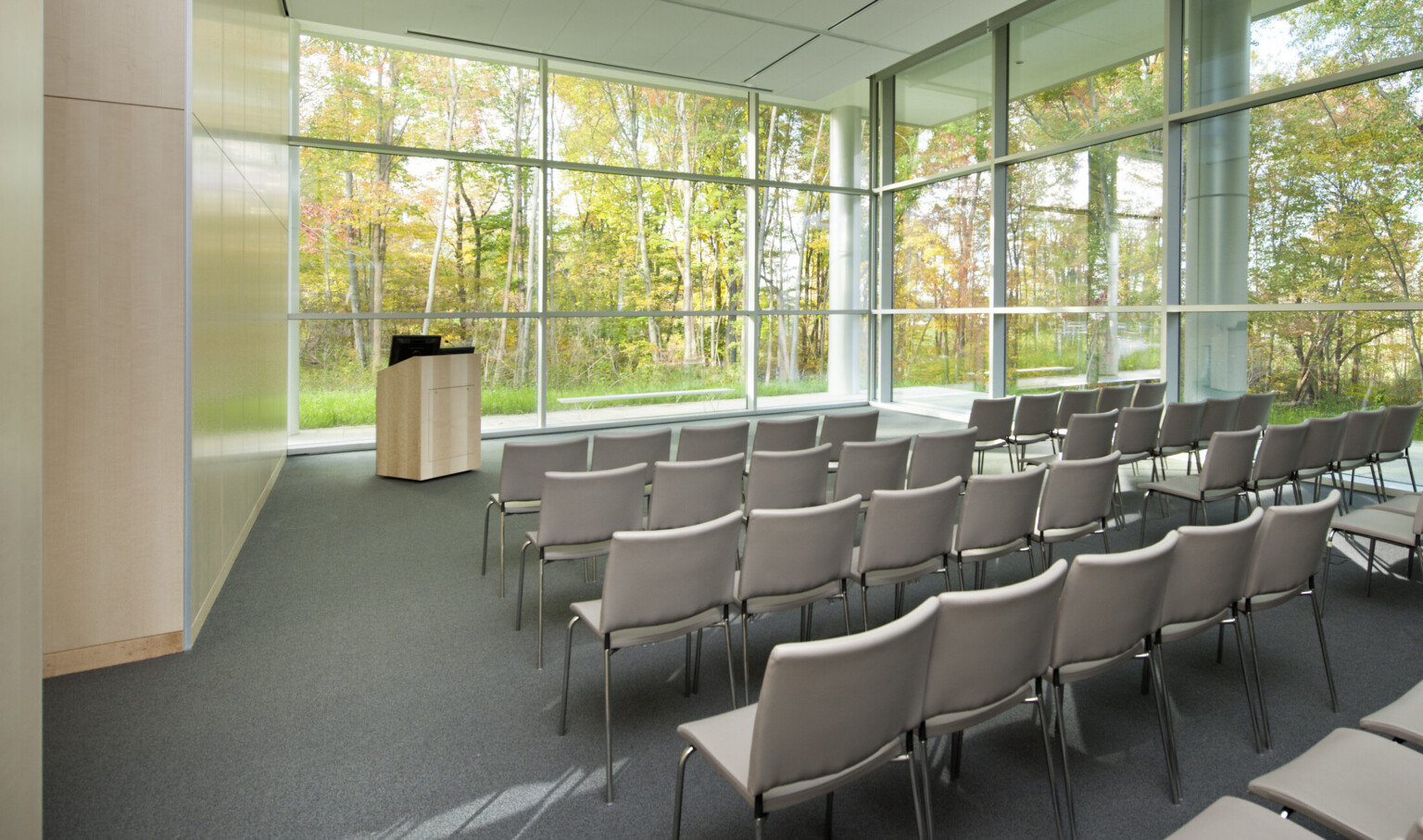 Auditorium with grey floor, wood left wall, and floor to ceiling windows on two walls with ground level view of trees.