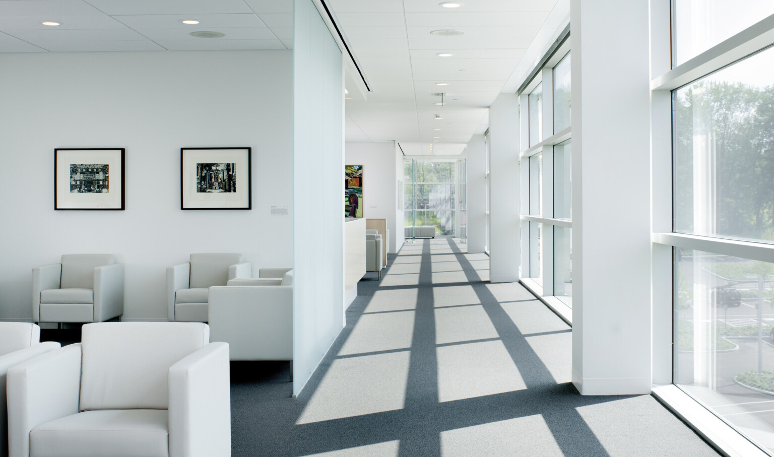 Hallway with floor to ceiling windows right and at end. Left divided into sections by white wall, each has seating areas
