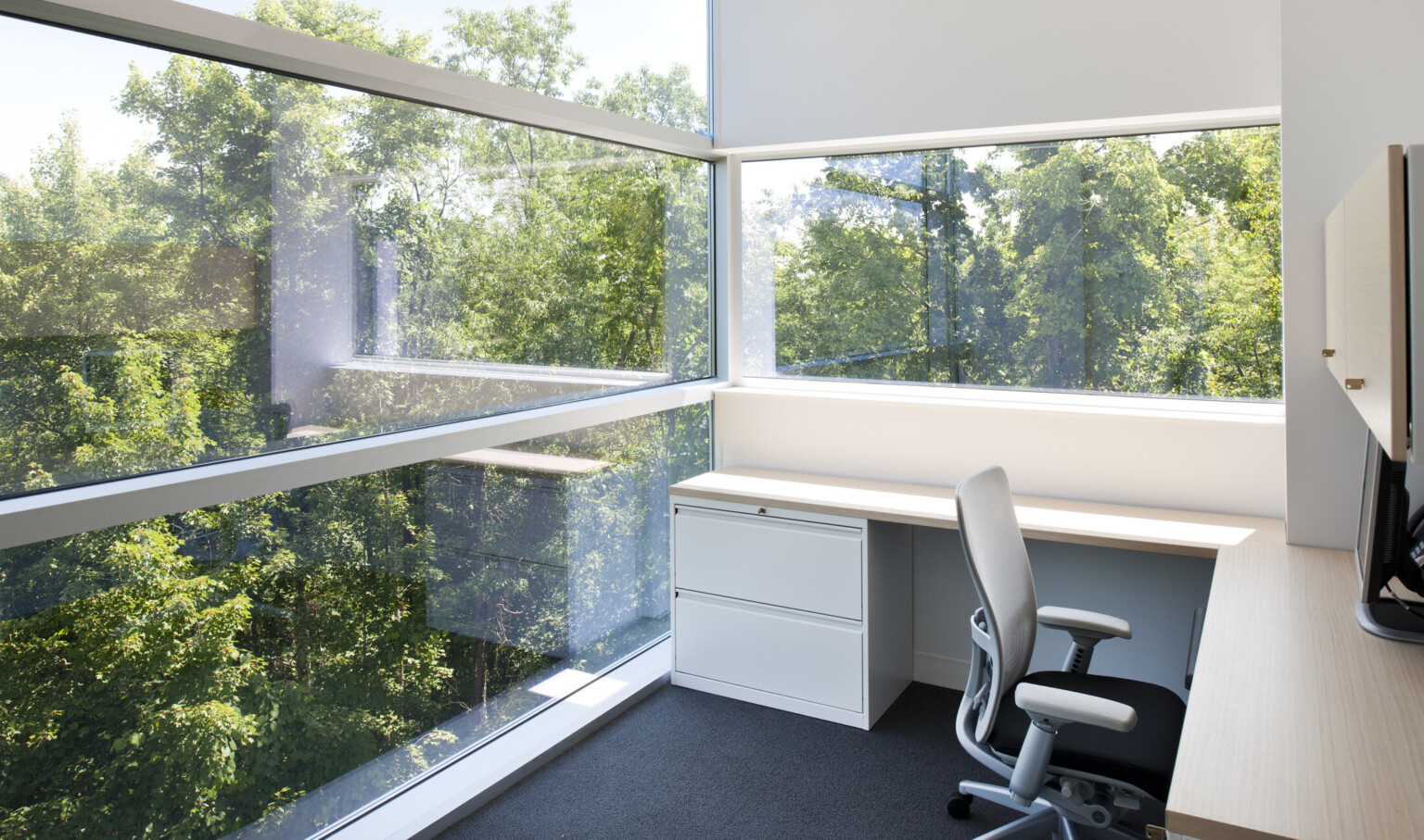 Corner view of office with L shaped desk and grey chair. Left is floor to ceiling window, back wall is white with wide window
