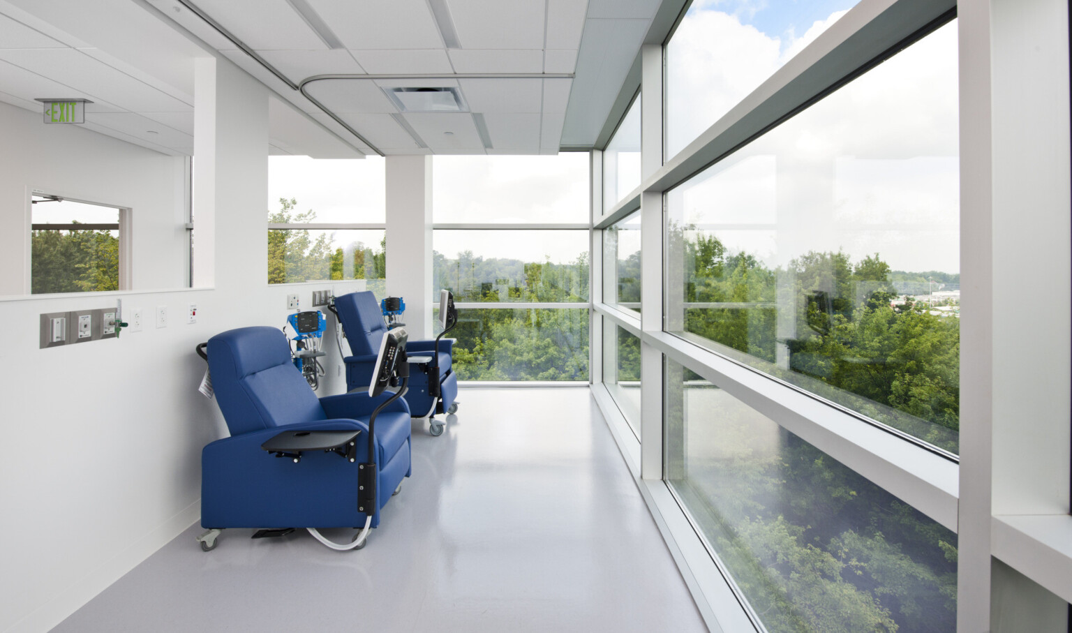 Blue chairs with attached table and screens next to half wall. Chairs face floor to ceiling window with view of trees