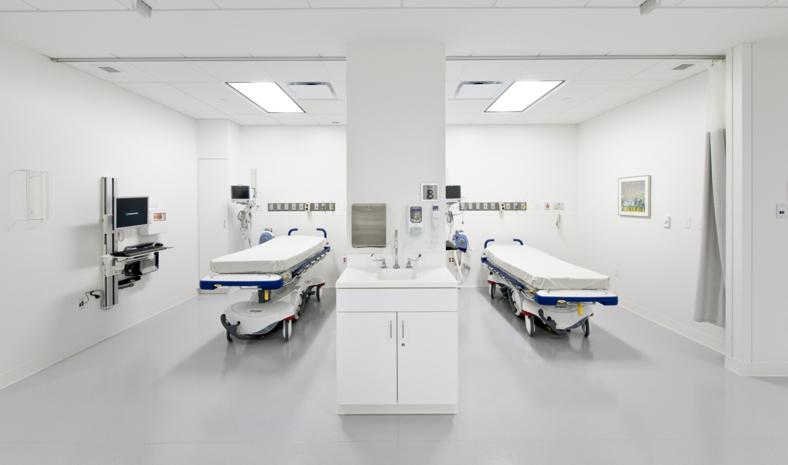 Two hospital beds in white room with equipment left. Desk and computer extend from left wall. White pillar center with faucet
