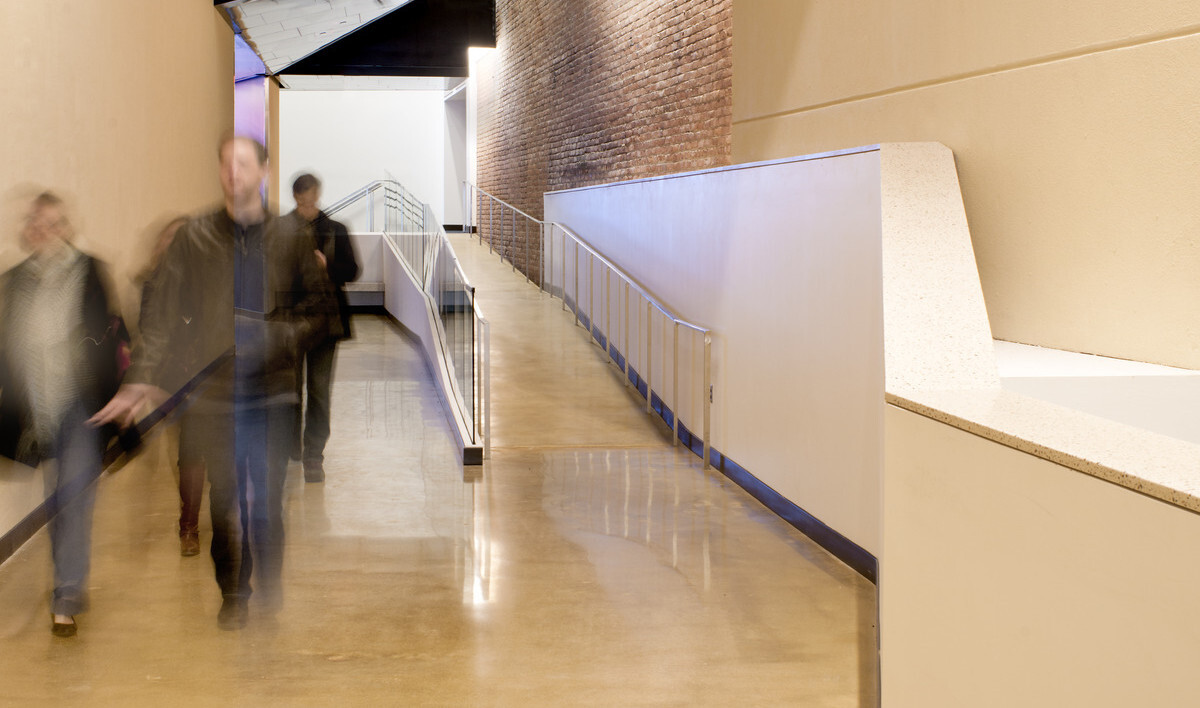 Hallway with ramp to the right, concrete yellow walls with exposed brick section reflected on metal ceiling section