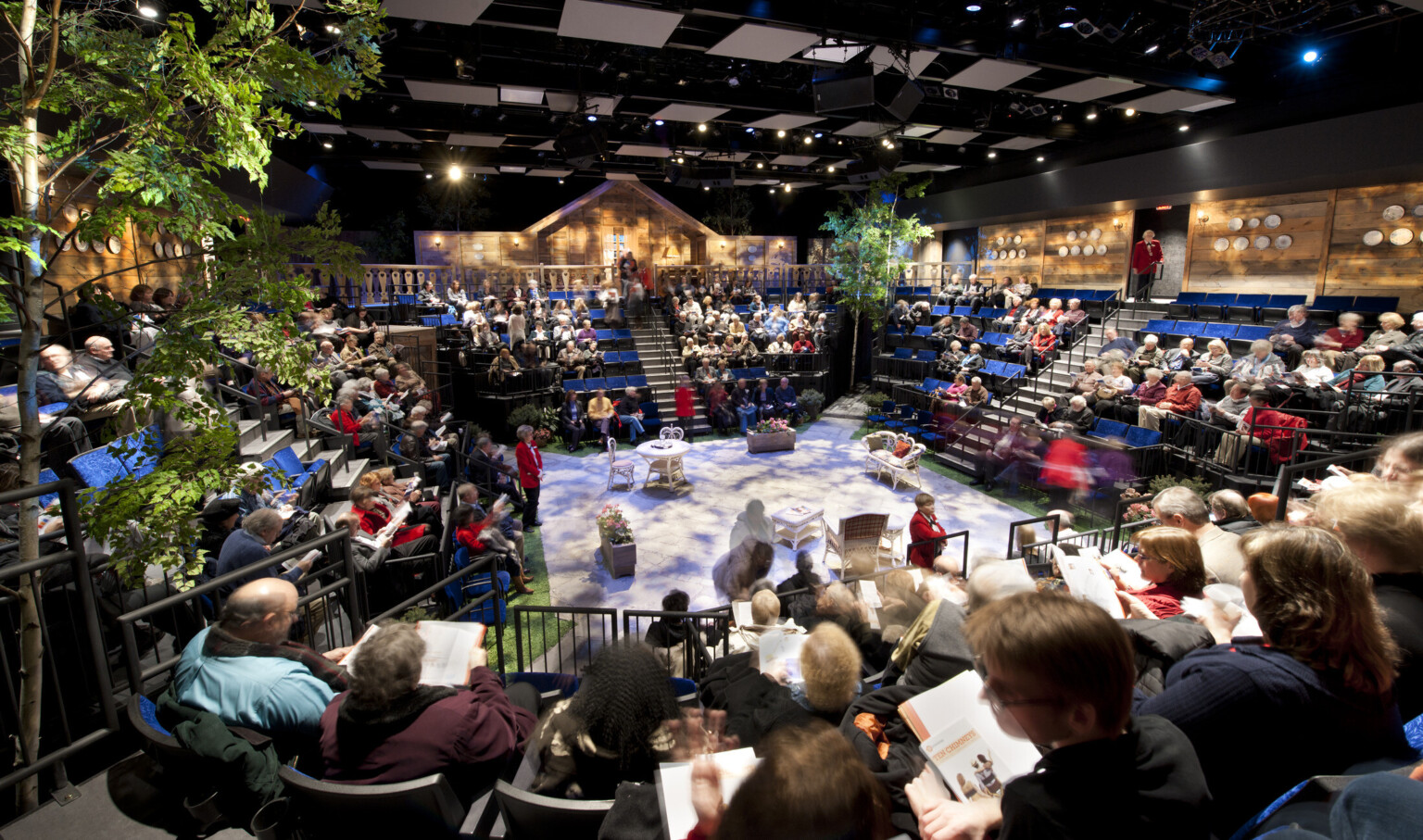 people seated at theater in the round with trees off a stage set with white table and chairs, textural lighting from above