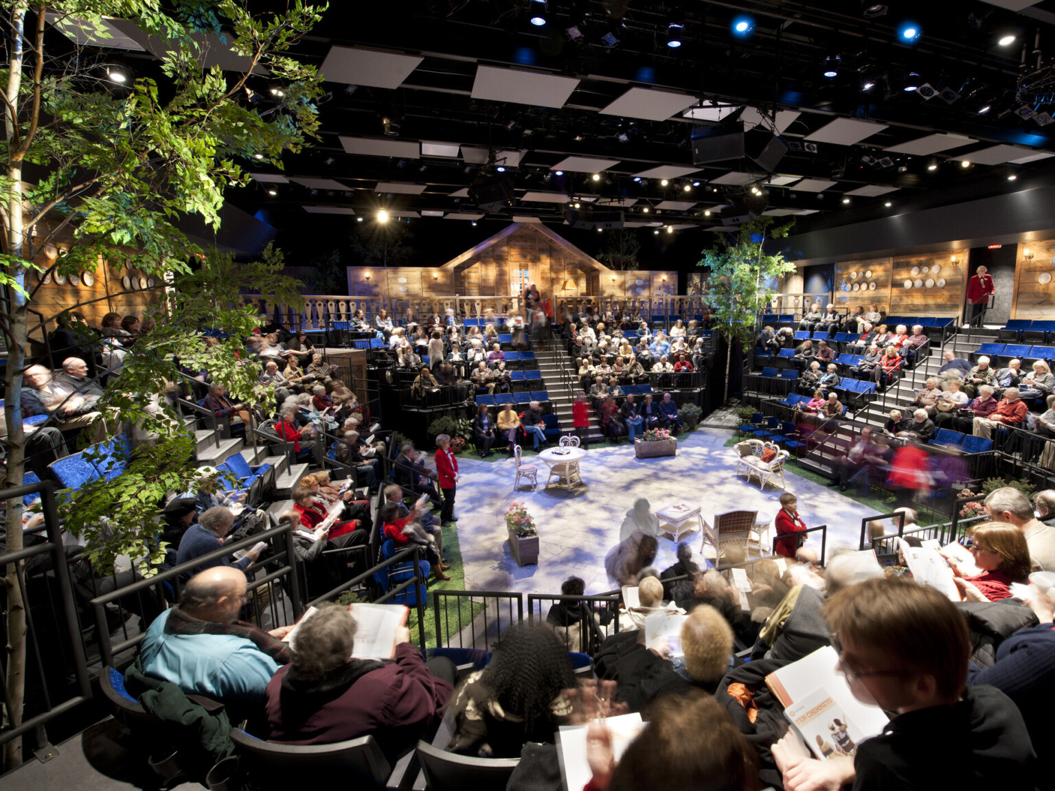 people seated at theater in the round with trees off a stage set with white table and chairs, textural lighting from above