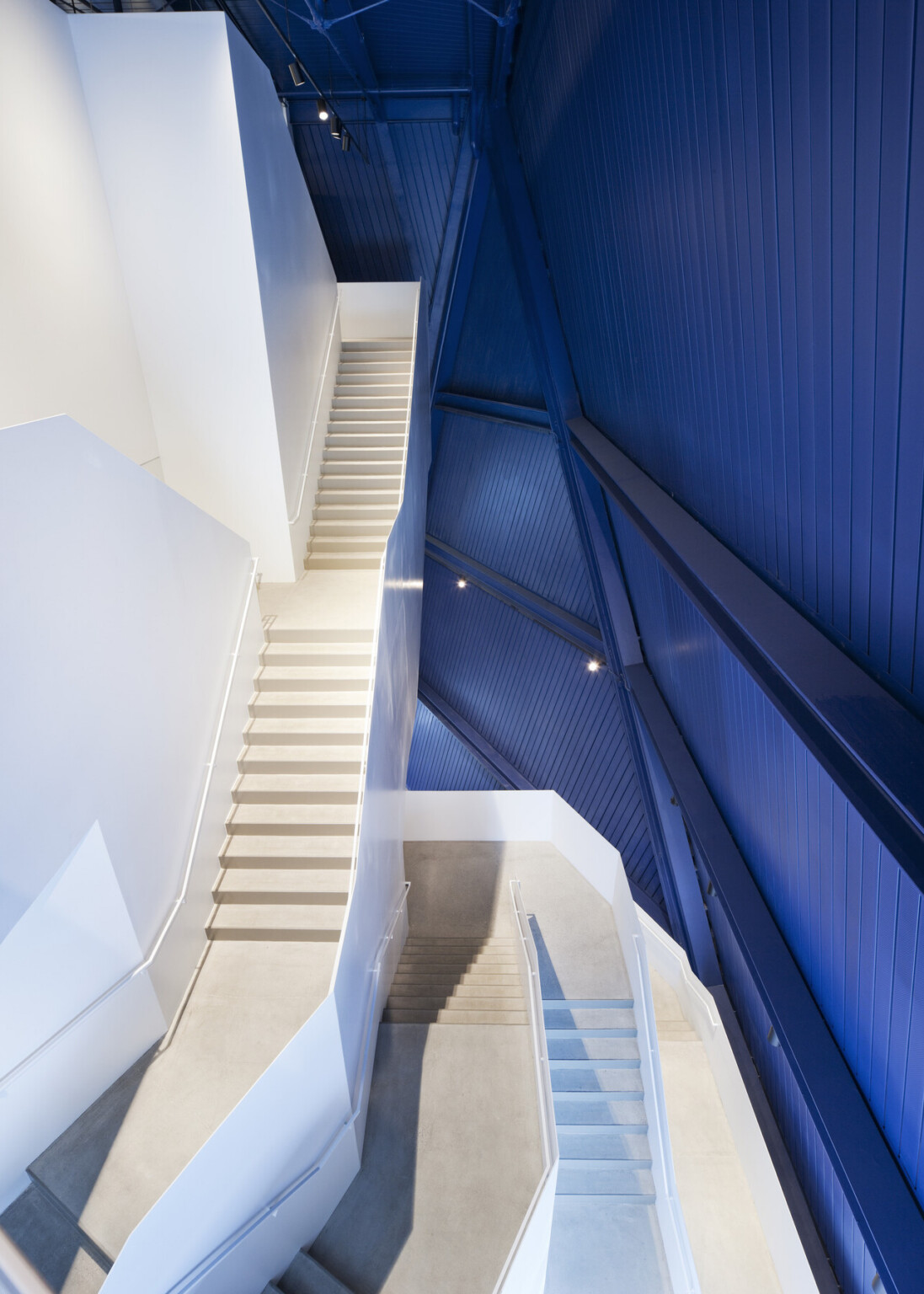 A modern interior staircase with clean white walls zigzags upward through a tall space. The staircases have light-colored steps and simple railings, while a dramatic angular wall on the right is clad in deep blue panels with visible structural framing. Overhead lighting highlights the sharp lines and geometric design, creating a contrast between the bright white surfaces and the bold blue wall.