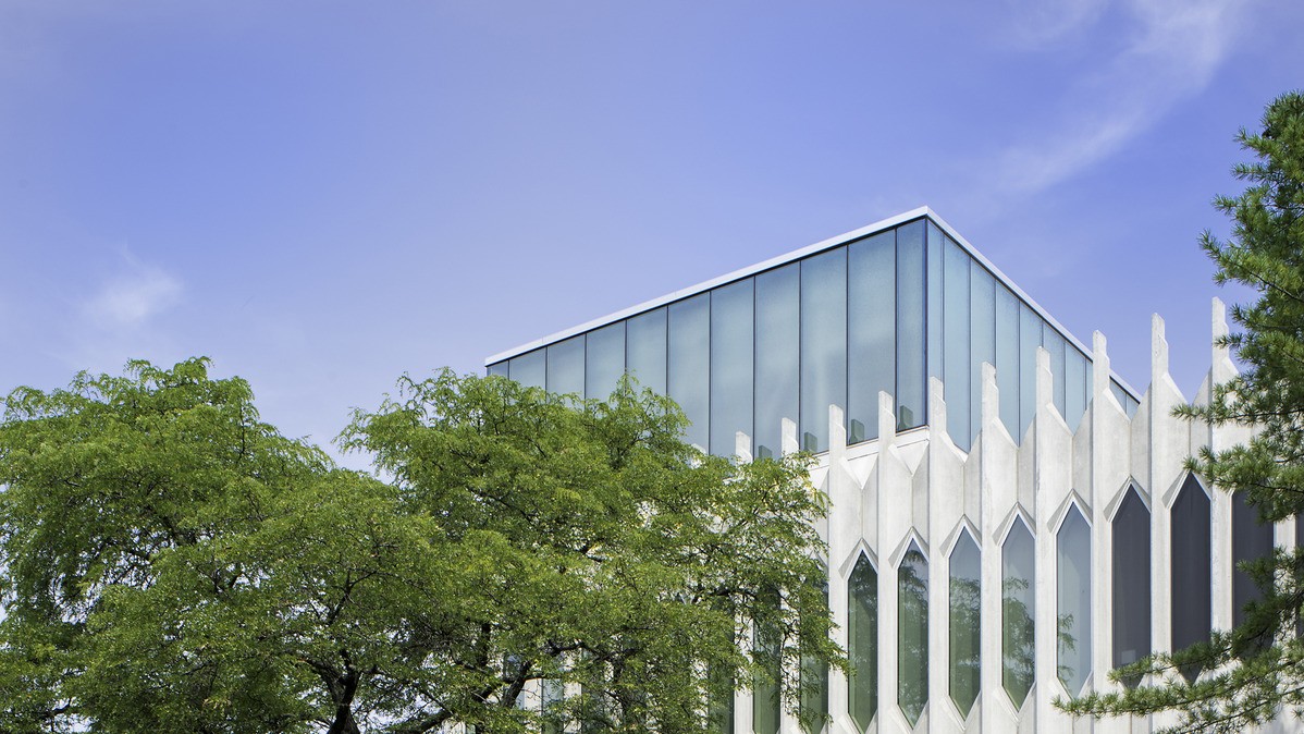 Cropped image of the top of a architecturally detailed building with trees in the foreground; white exposed steel structure in front of glass building