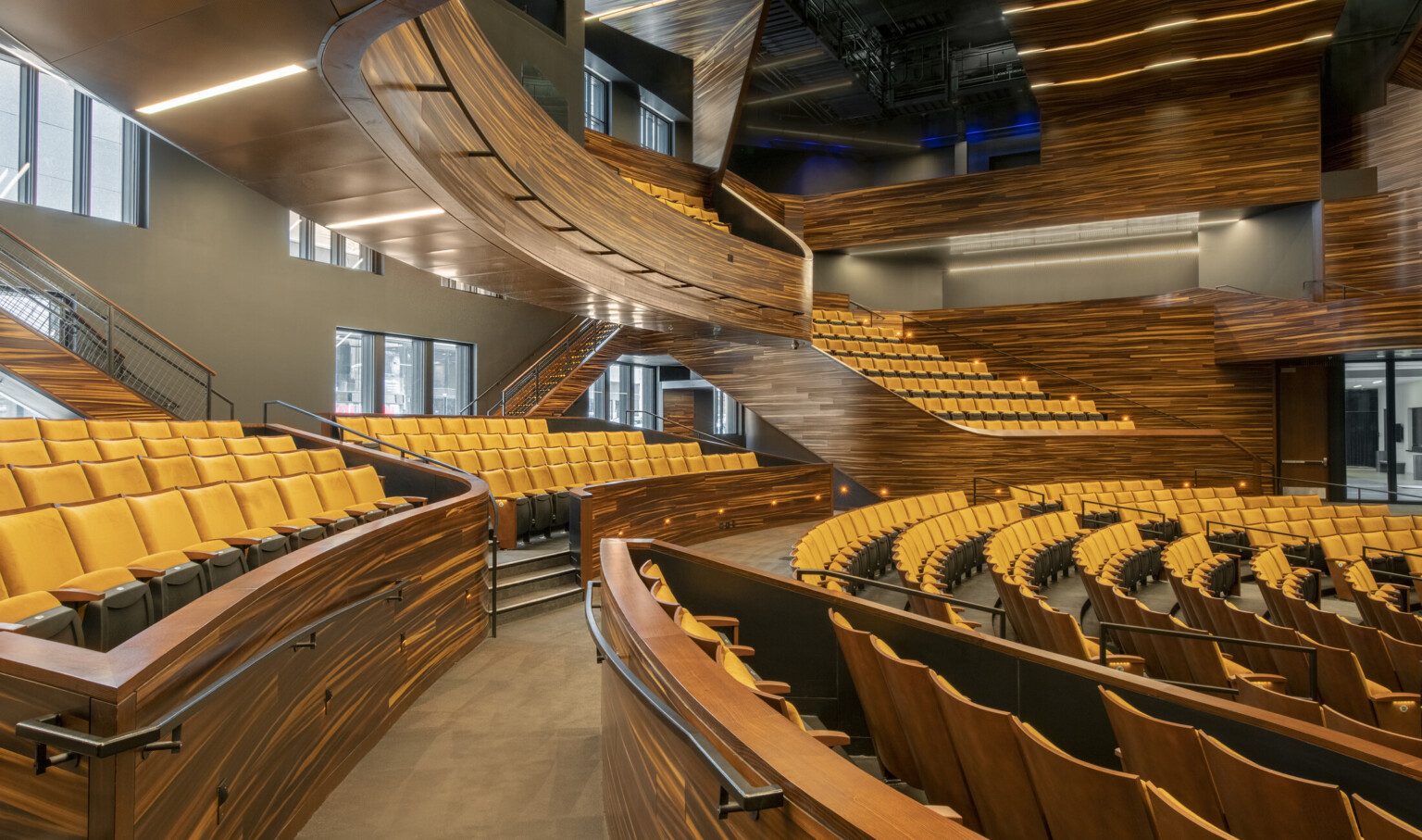 Stadium style yellow seating and abstract stripe stained wood details, balcony, and bannisters with recessed aisle lighting