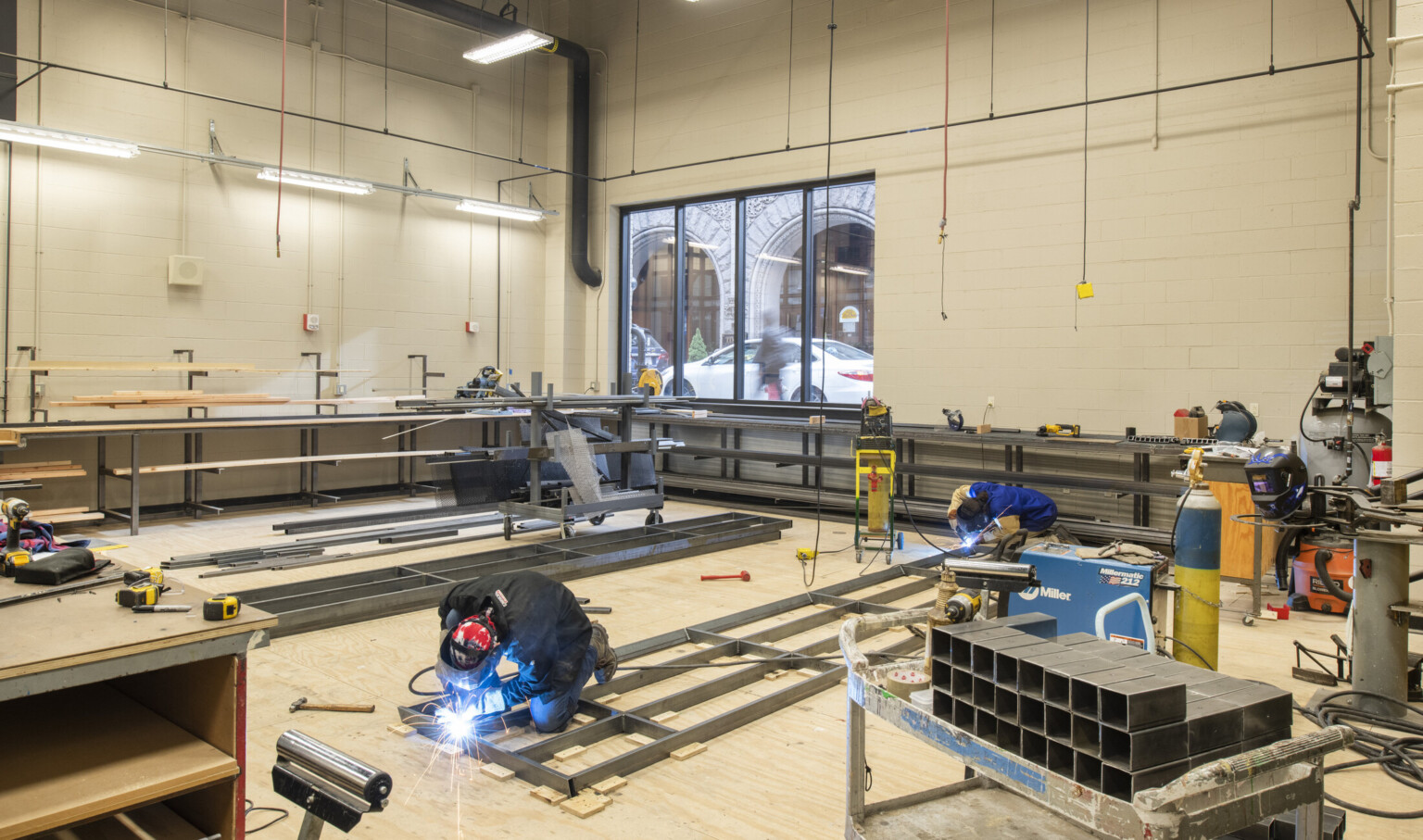 Workspace with welding equipment and tools, metal is being worked. A large window to the back looks out on street level
