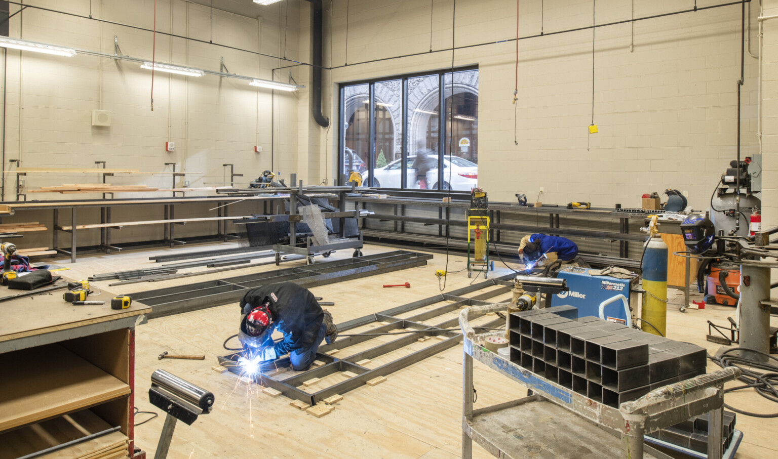 Workspace with welding equipment and tools, metal is being worked. A large window to the back looks out on street level