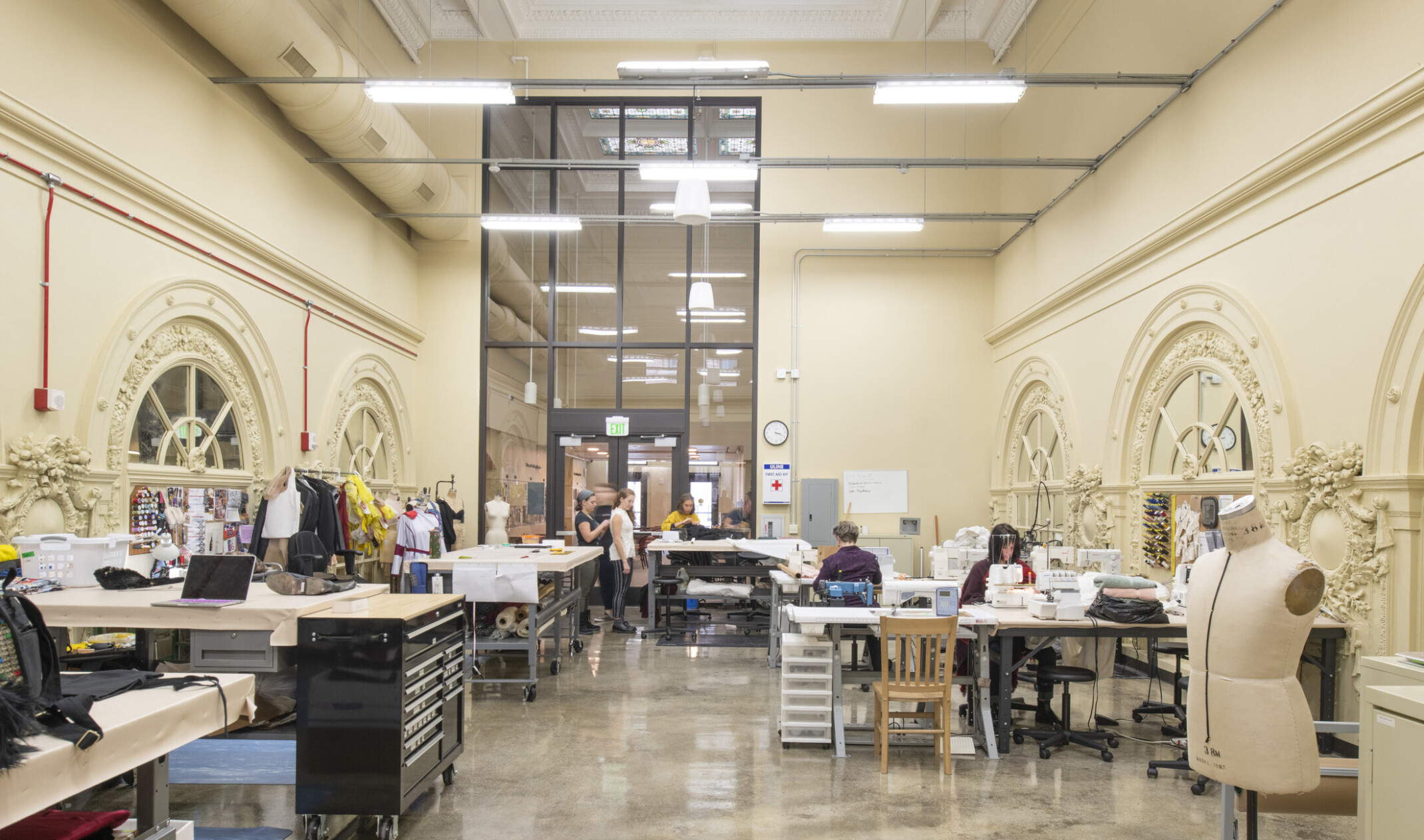 Double height long room with sewing machines and arched mirrors with ornate molding details on either side of work tables
