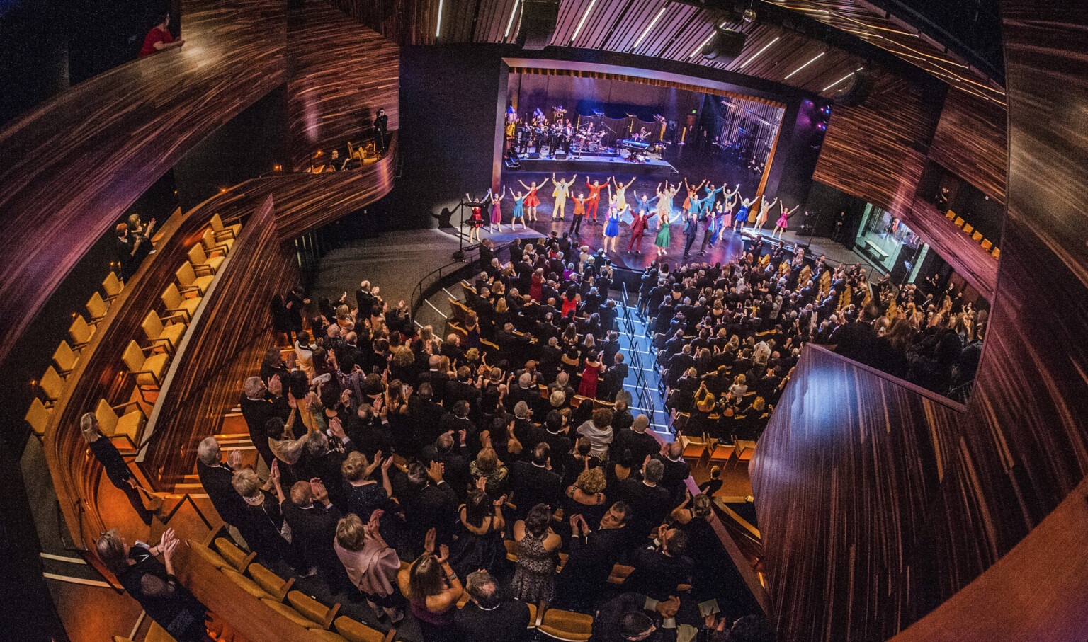 fisheye view of theater with audience and cast bowing on stage, surrounded by dark wood paneling and deep purple lights