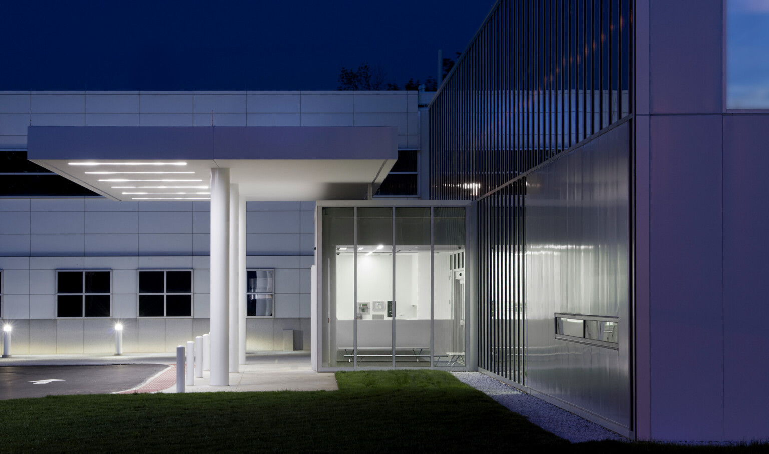 Side view of entry with glass vestibule with benches. Overhang above entry to driveway. Grey wall behind with square windows