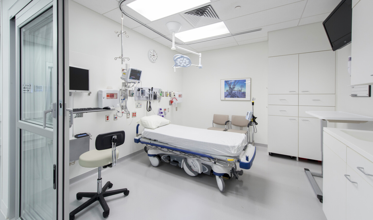 Hospital bed in white room with equipment over head. Two beige chairs behind bed next to 2 large cupboards. Desk on left wall