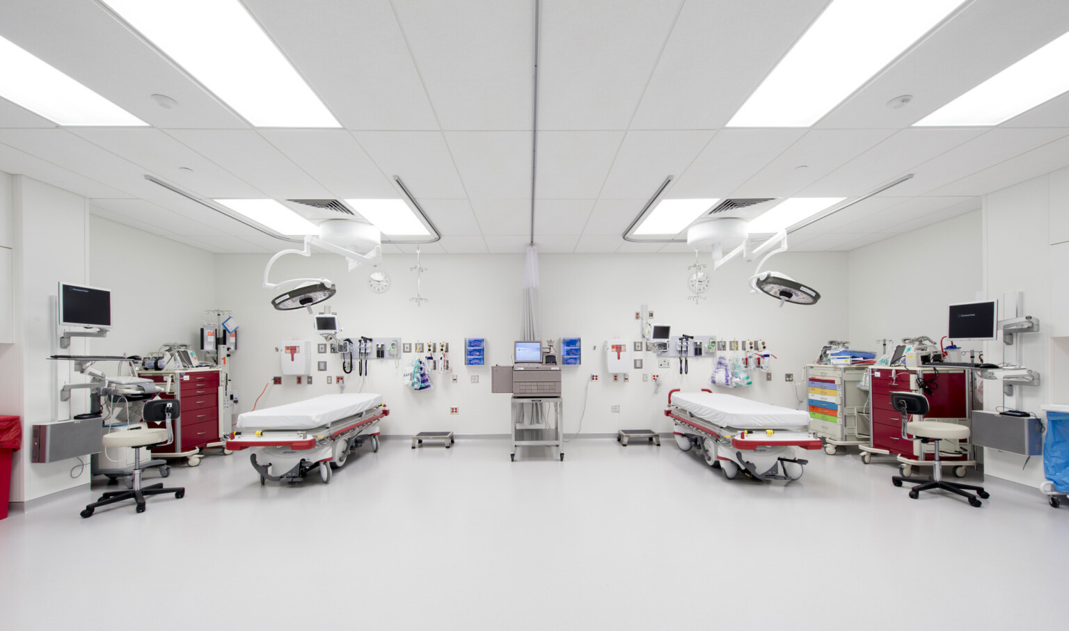 White room with 2 hospital beds with large circular whites above. Machine on rolling table center. Medical carts on each side