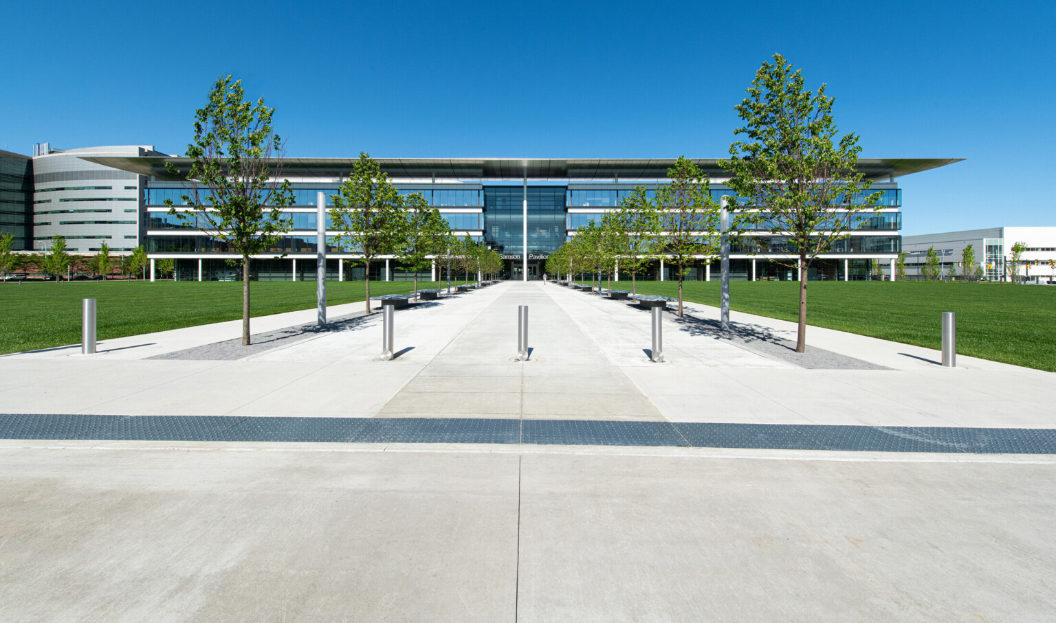 Building front with 4 stories of floor to ceiling windows and recessed double height window entry. Tree lined walkway to door