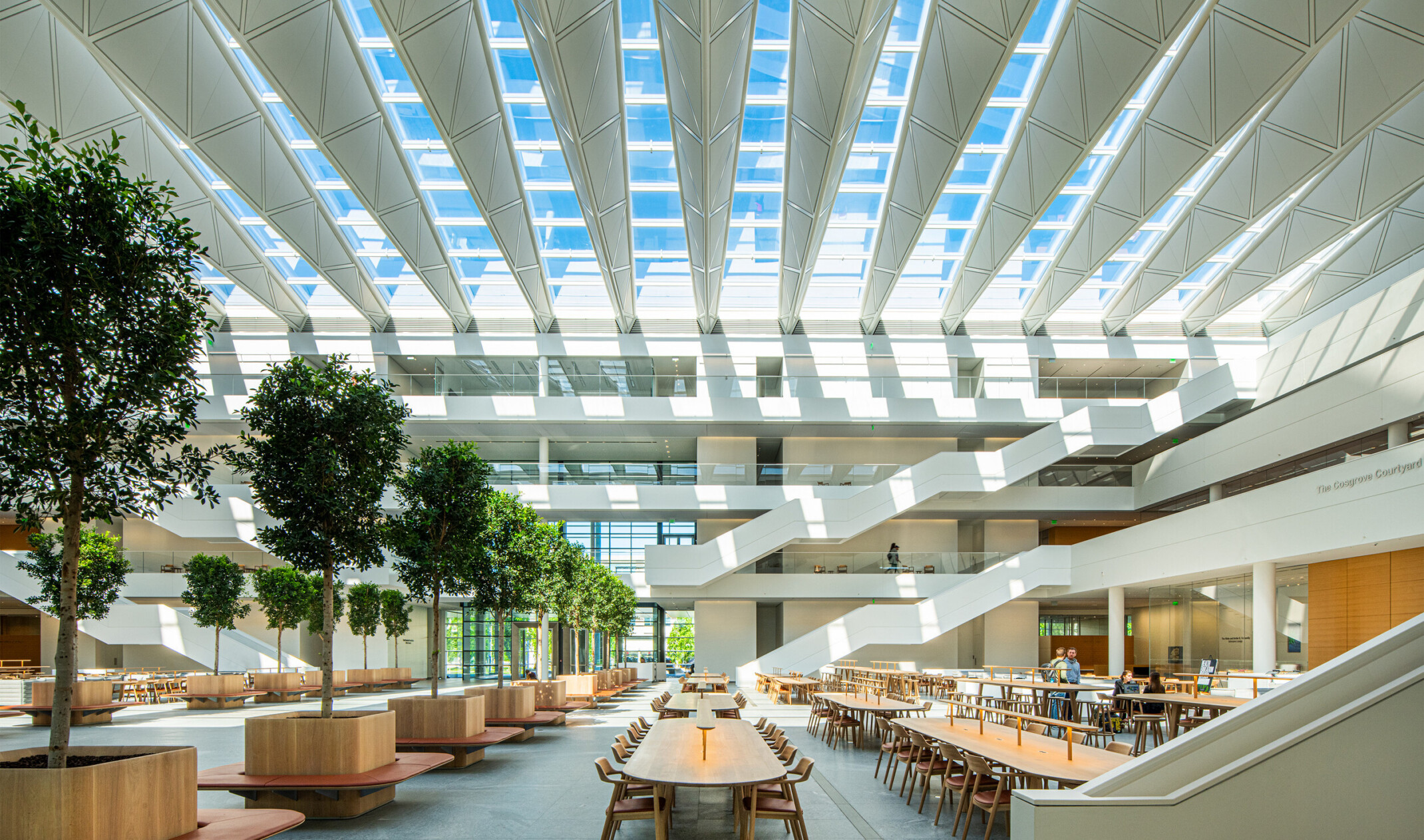 Atrium seen from ground level. Long tables with chairs on either side of tree lined walkway. White angular staircases at back