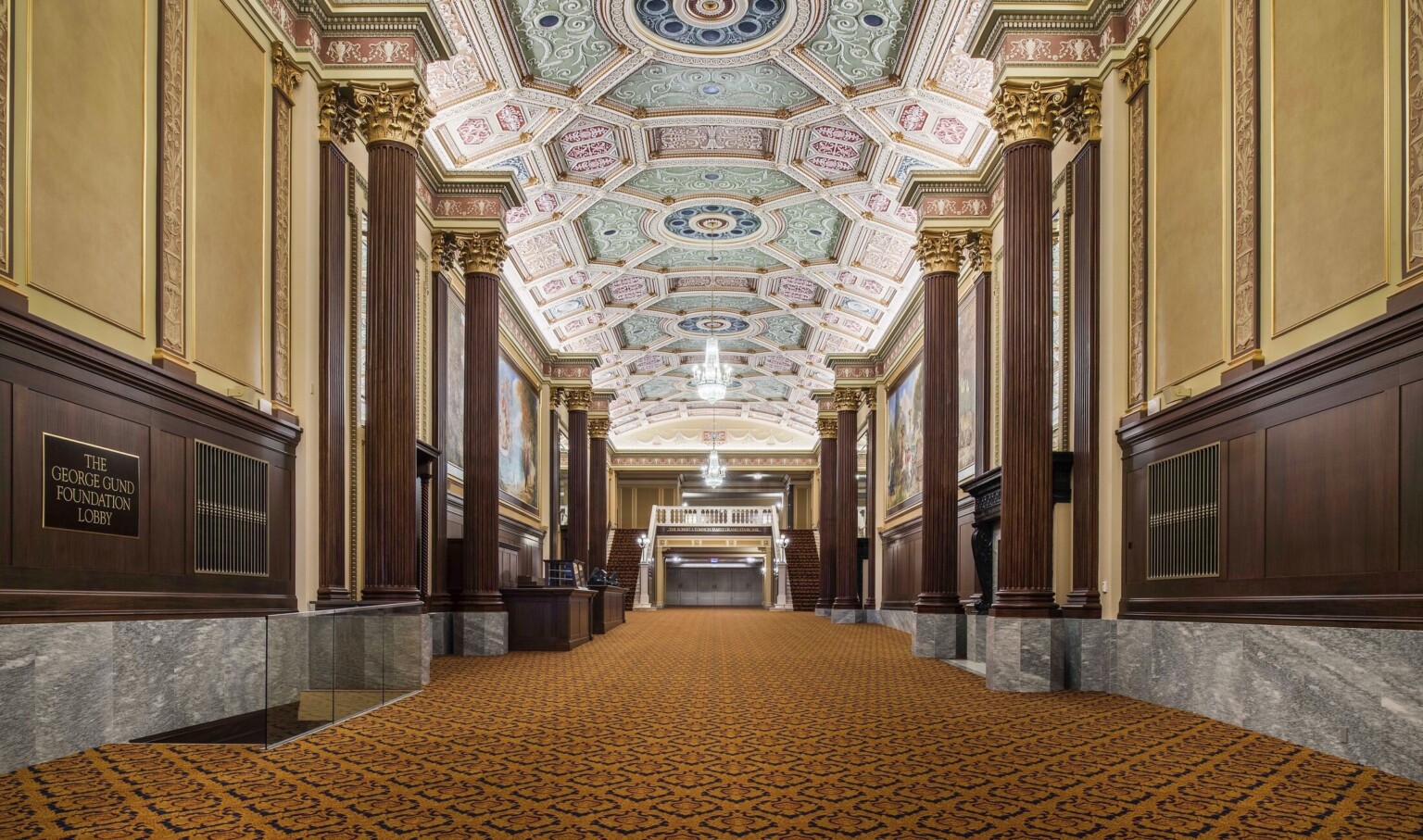 Grand staircase carpeted beneath ornate multicolored coffered ceiling with chandelier framed by ribbed pillars