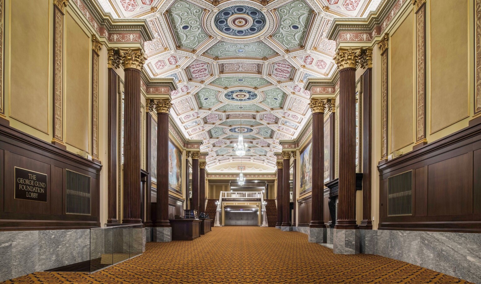 Grand staircase carpeted beneath ornate multicolored coffered ceiling with chandelier framed by ribbed pillars