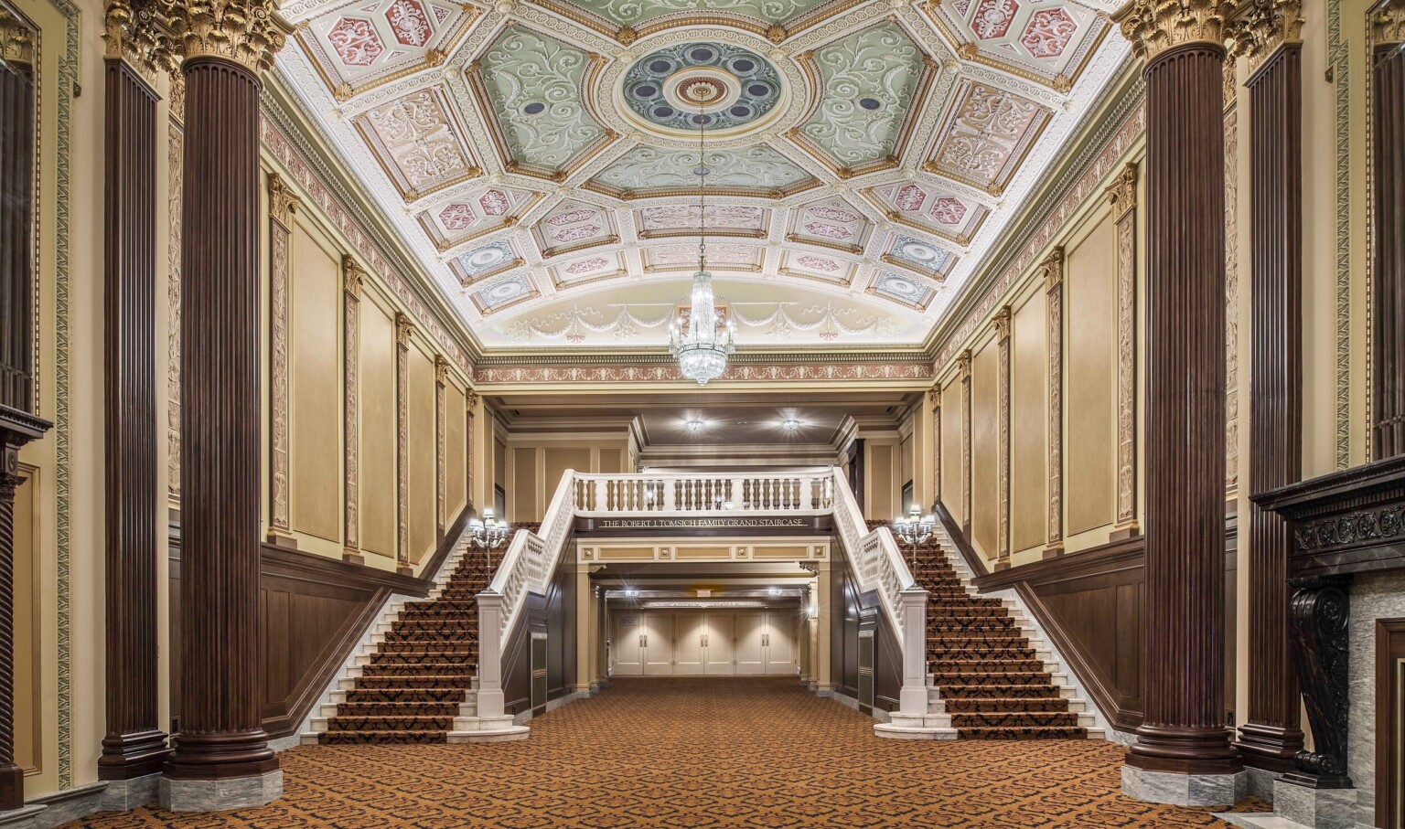 Grand staircase carpeted beneath ornate multicolored coffered ceiling with chandelier framed by ribbed pillars