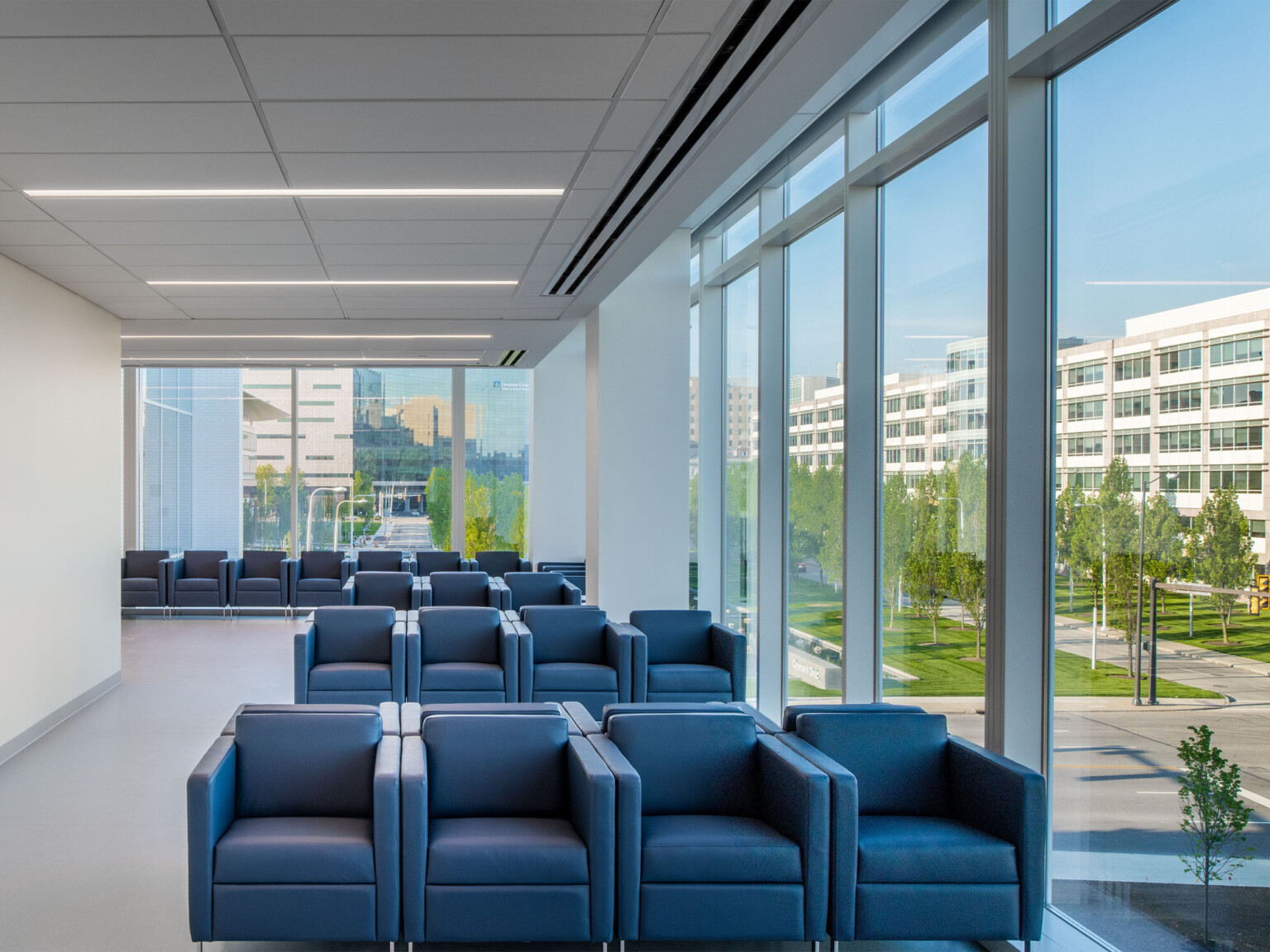 Seating area with blue arm chairs in white room with two walls of full length windows. Recessed lines of light from above