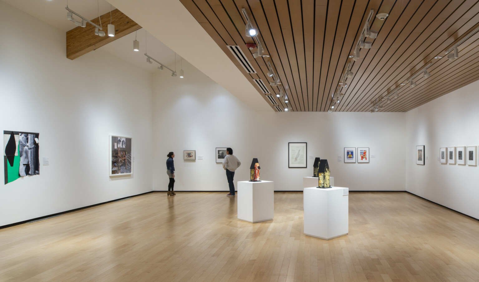 culture seekers view exhibit in performing arts center, timber paneled ceilings, overhead spotlights, gleaming hardwood floors, high-ceilings, hanging art on white walls, sculptures on modern pedestals of various sizes