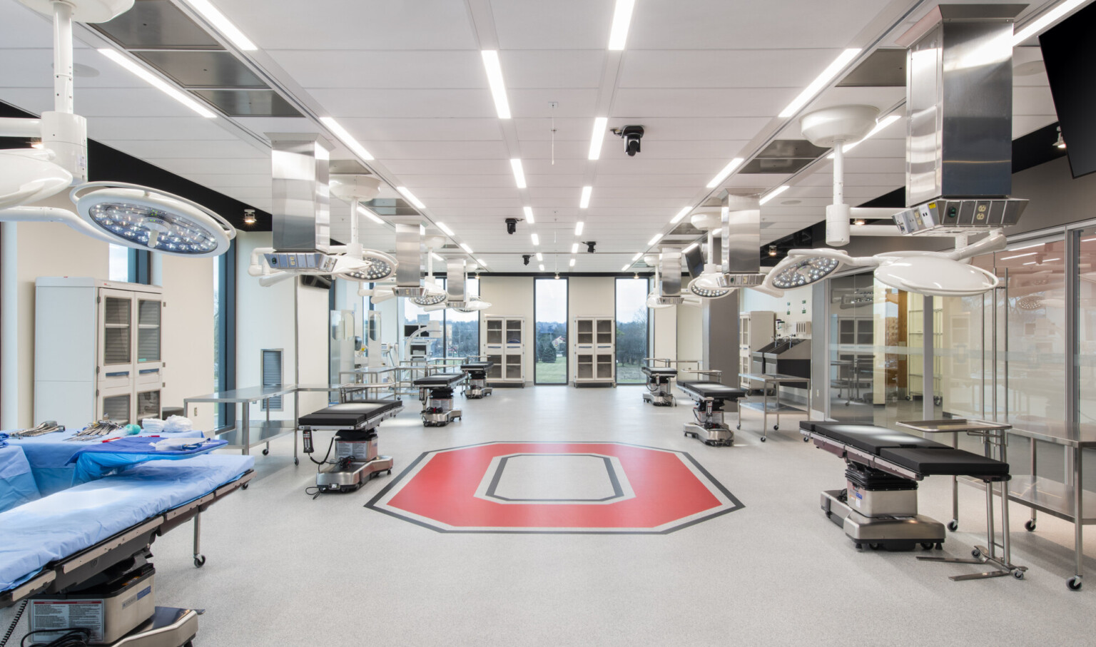 Inside view of room with 6 hospital beds with 2 circular lights hung above, tables next to the bed and red O on floor