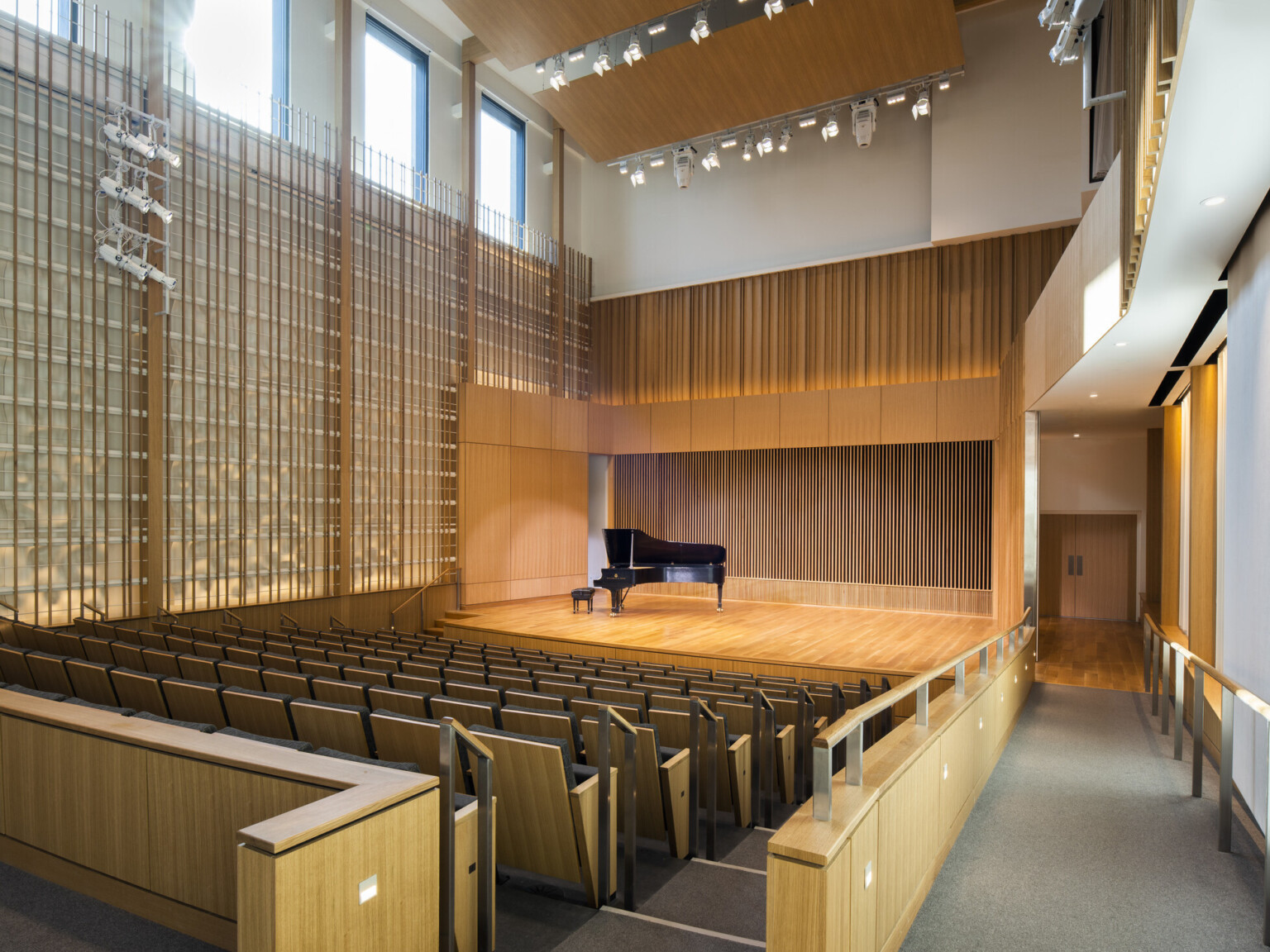 Angled view from side aisle of same recital room with piano, empty, illuminated with stage by theater lighting hanging from above