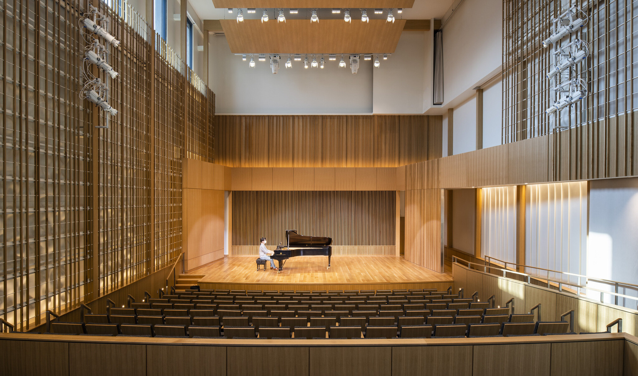 Man sitting at piano in double height recital room auditorium with theater seating. Wood panel designs on walls and wood acoustic panel
