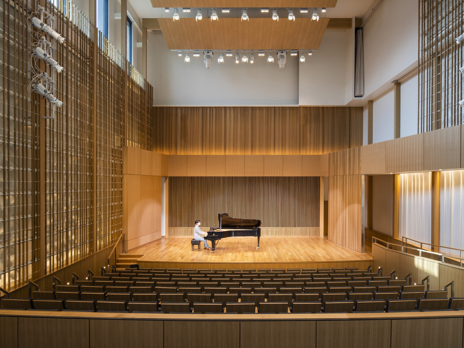 Man sitting at piano in double height recital room auditorium with theater seating. Wood panel designs on walls and wood acoustic panel