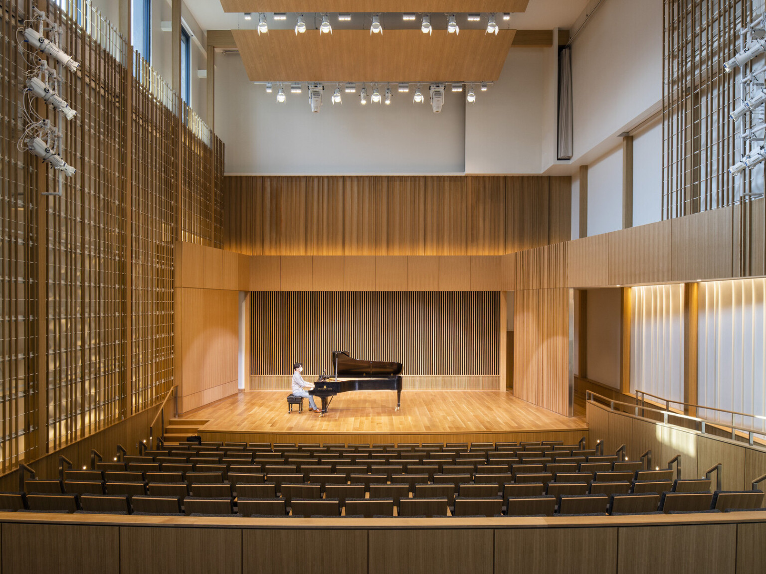 Man sitting at piano in double height recital room auditorium with theater seating. Wood panel designs on walls and wood acoustic panel